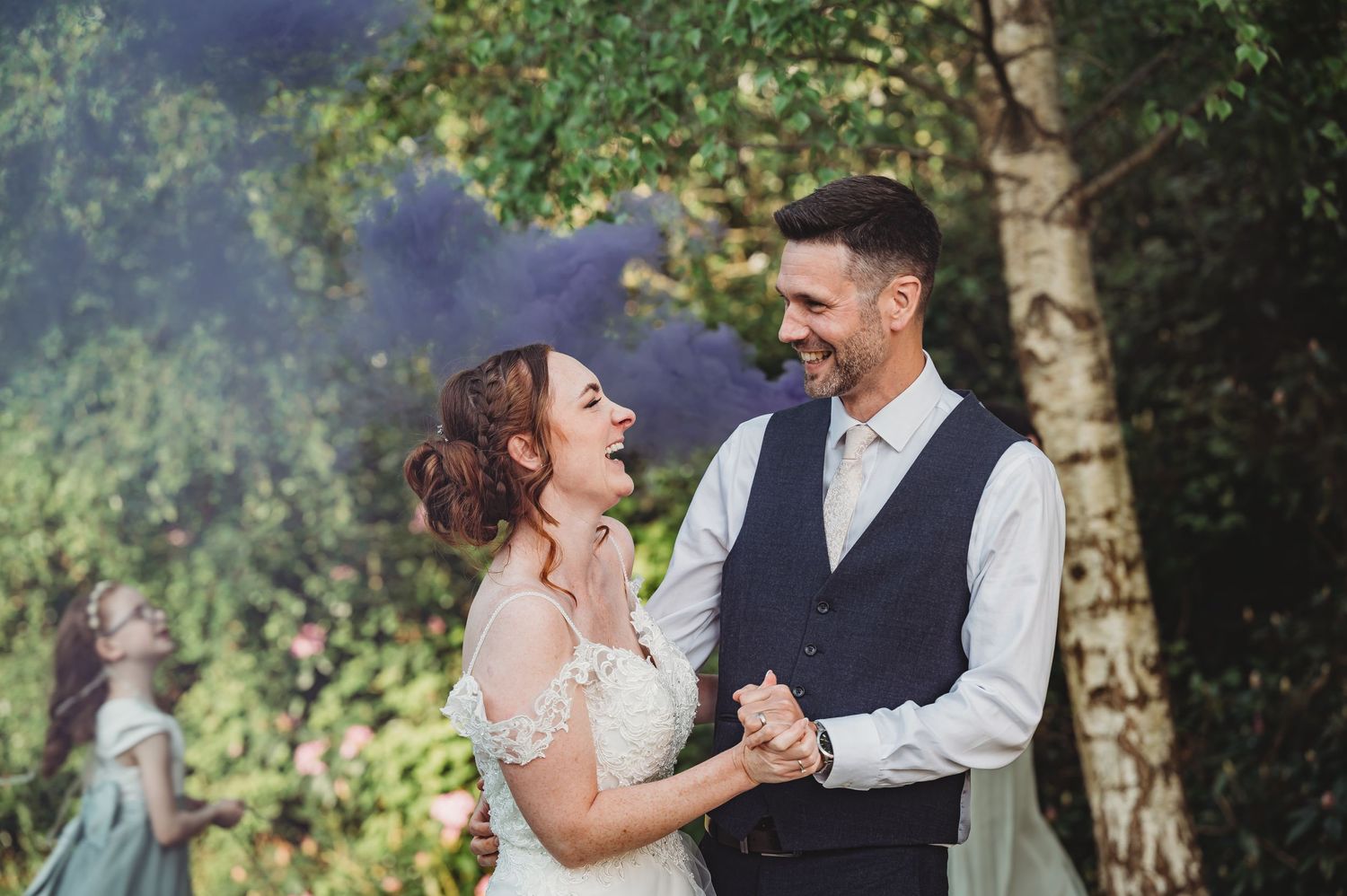A wedding couple shares a joyful laugh together in a scenic outdoor setting with trees in the background.