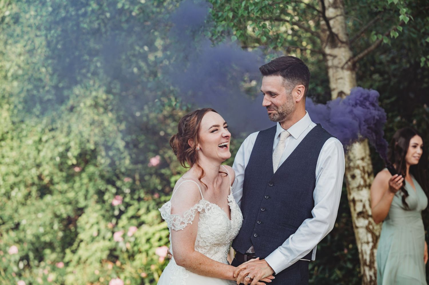 A joyful bride and groom share a candid moment together outdoors surrounded by purple smoke and greenery.