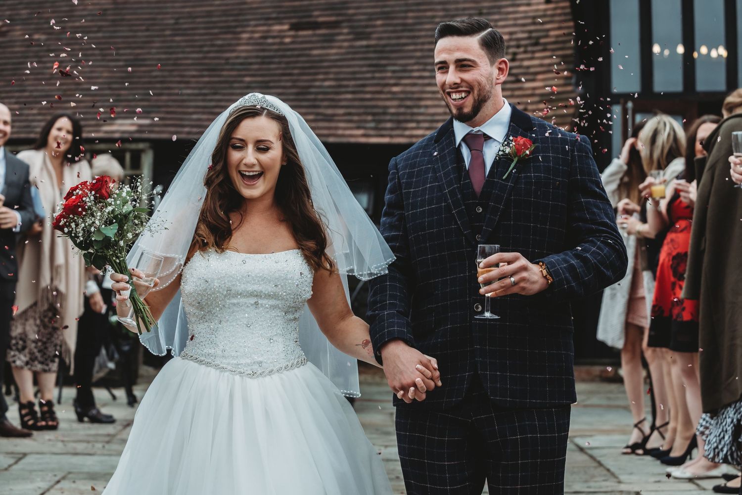 A joyful celebration with a white wedding gown and plaid suit as guests gather outside a brick building.