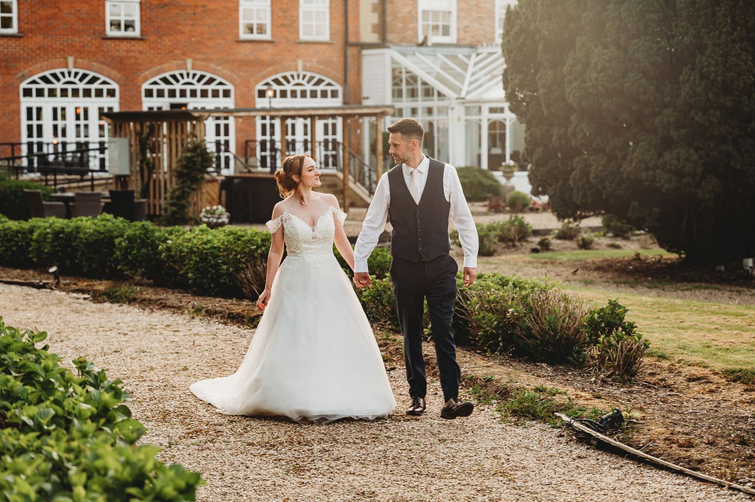 Newlyweds walk hand in hand along a garden path in front of a stately brick manor house on a sunny day.