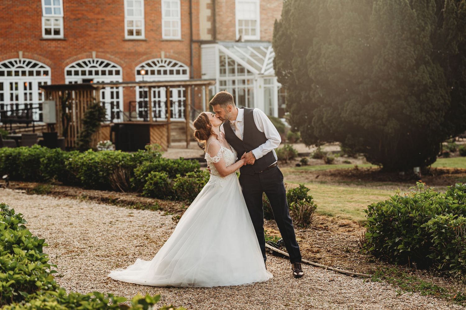 A romantic wedding couple embraces on a gravel path outside a historic red brick mansion with ornate windows.