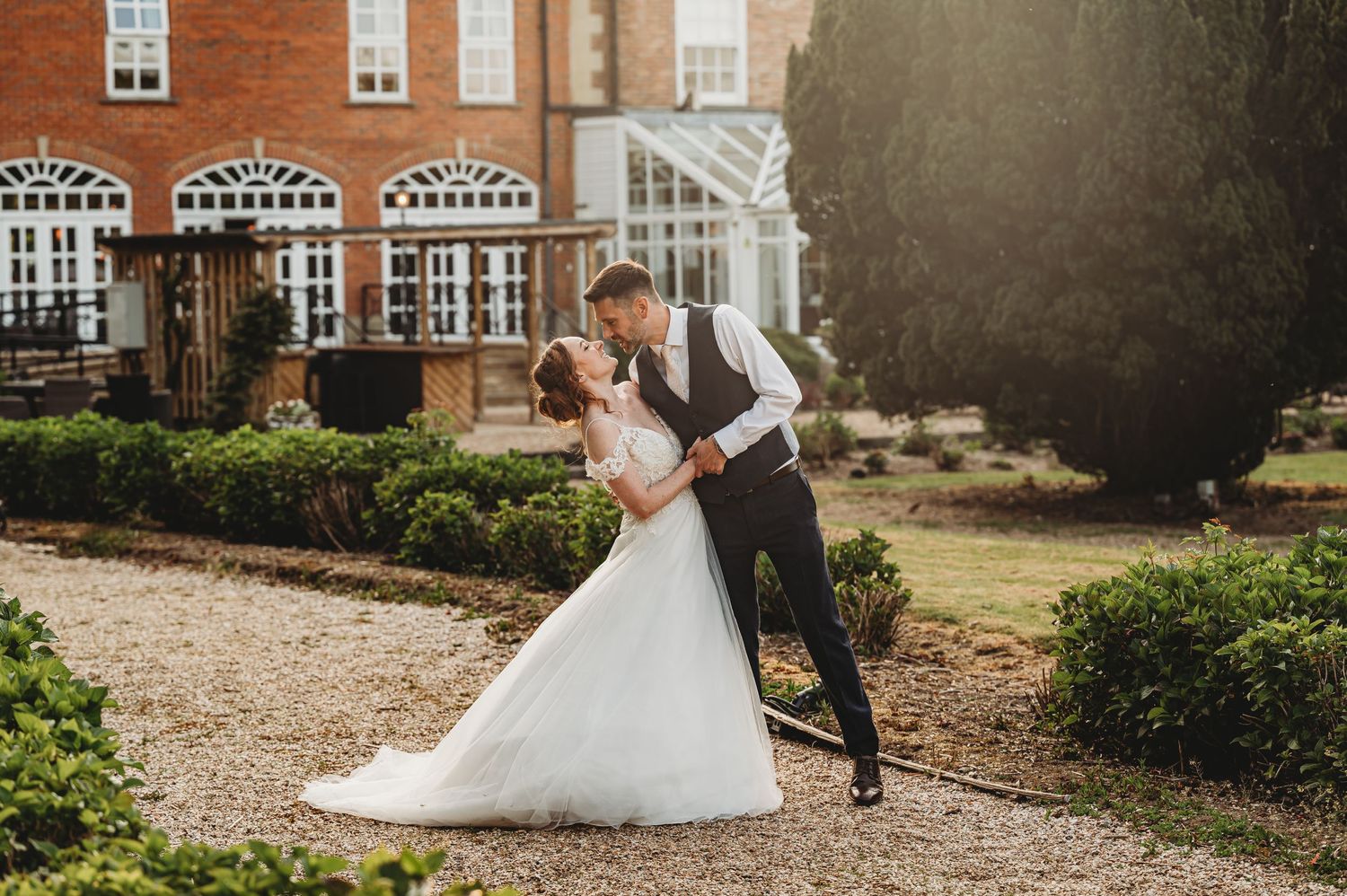 A romantic wedding couple embraces outside a grand red brick mansion with ornate windows and sunlit gardens.