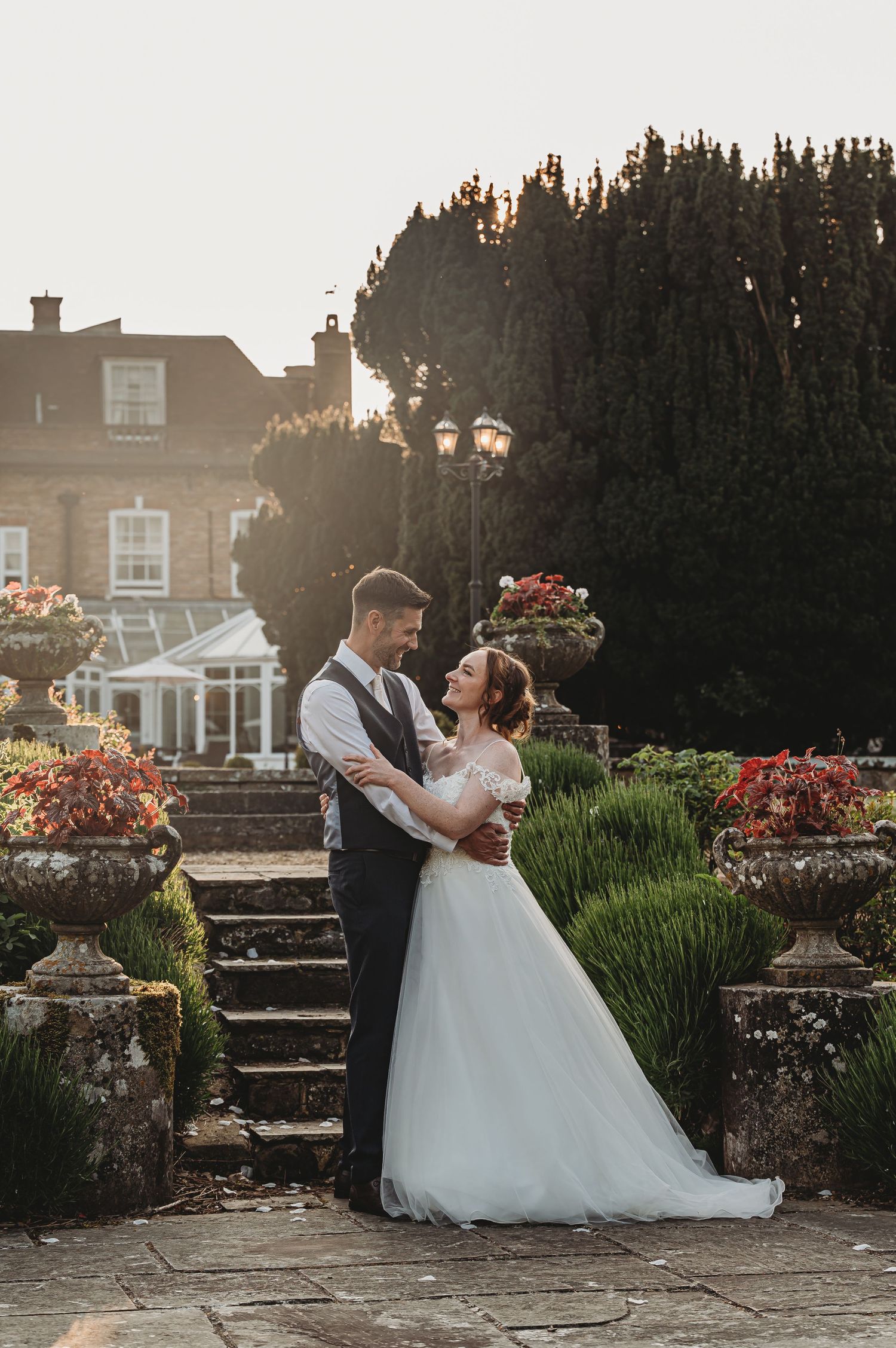 A couple embraces on stone steps at sunset in front of a classic country house with blooming gardens.