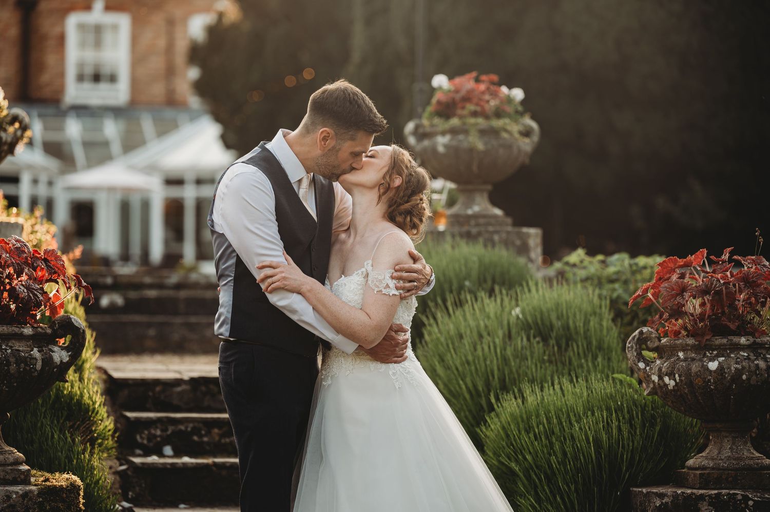 A romantic wedding couple embraces on garden steps surrounded by flowers and ornate stonework at sunset.