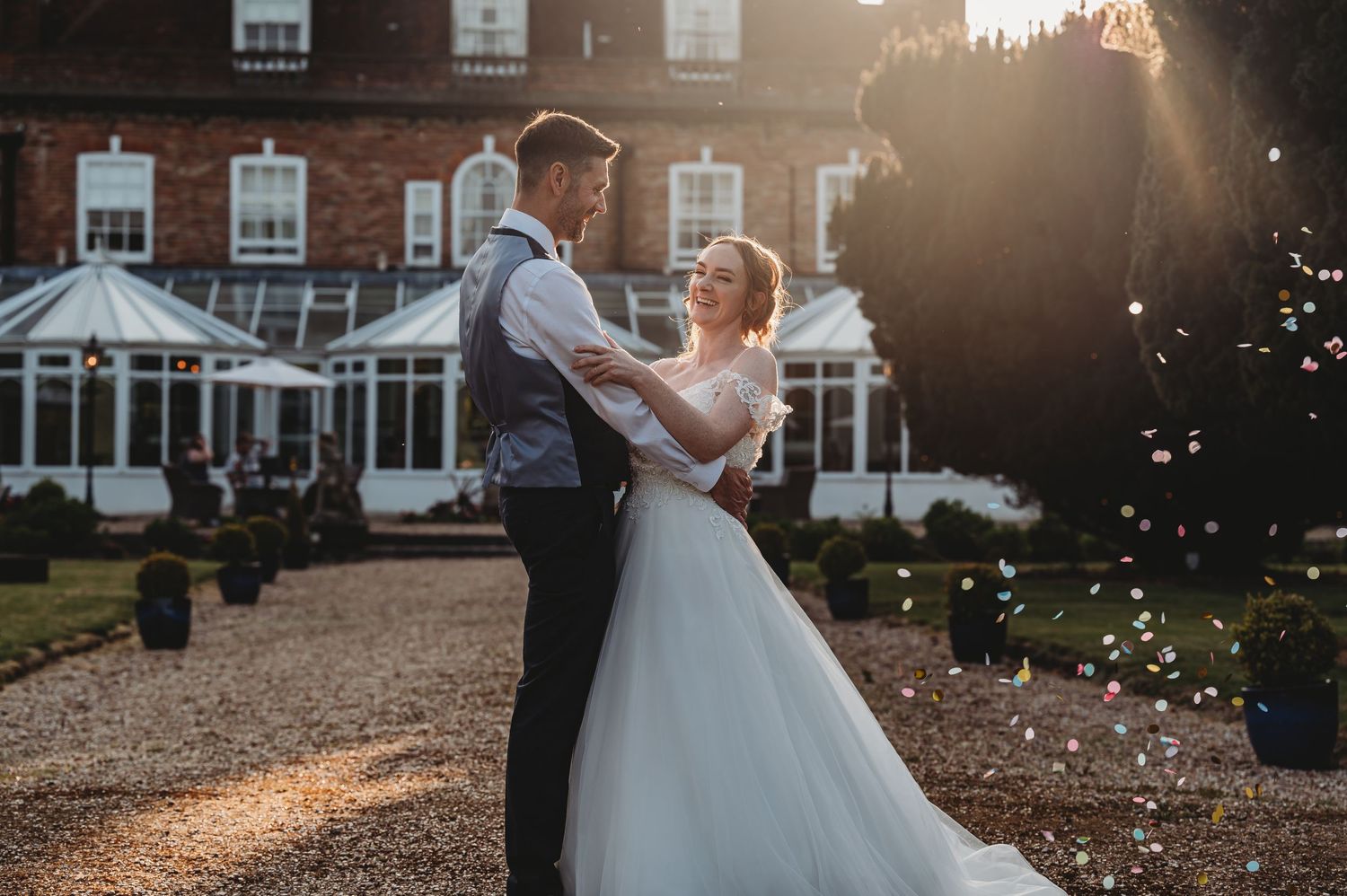 A newlywed couple dancing at sunset in front of a grand brick estate with golden light and floating bubbles.