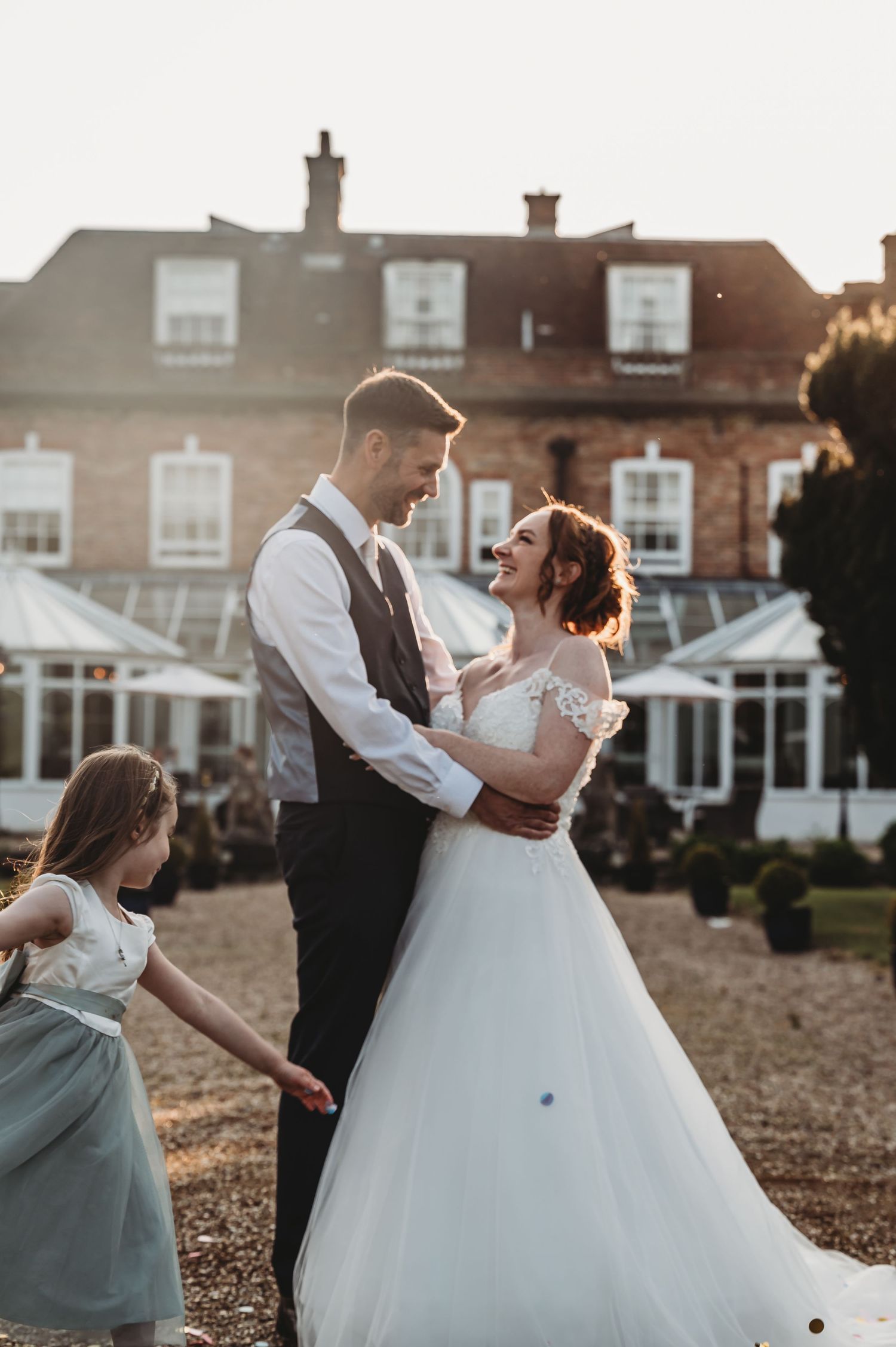 Wedding couple embraces in front of large brick mansion with conservatory as young bridesmaid in blue dress looks on.