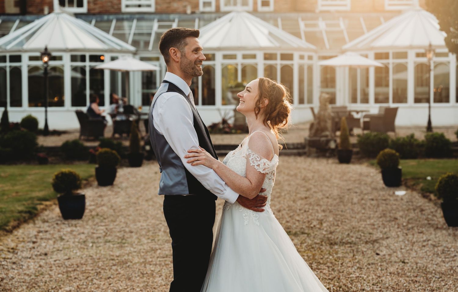 Romantic couple embraces on gravel path in front of elegant conservatory venue at golden hour sunset.