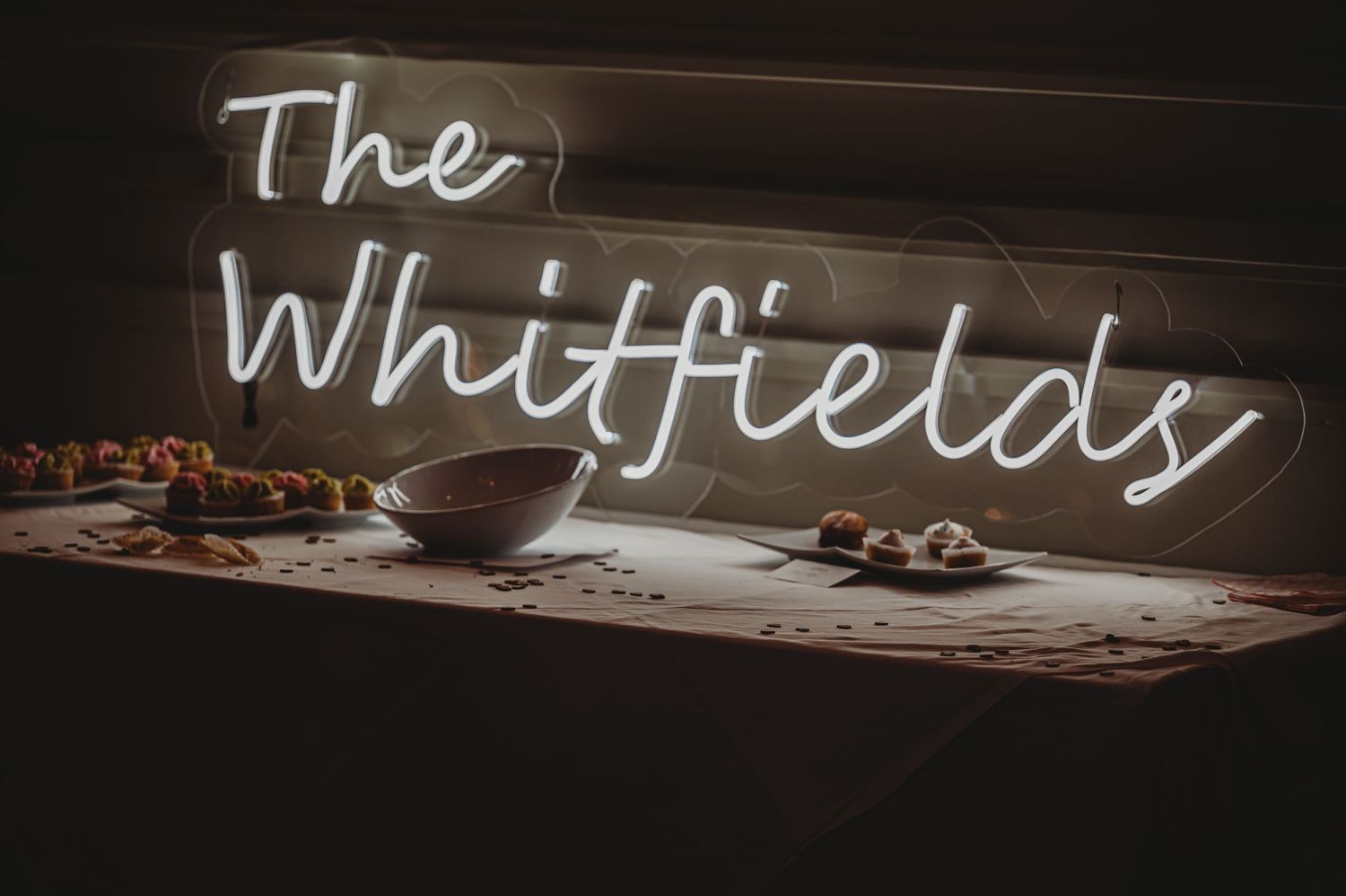 A glowing white neon sign reading 'The Whitfields' illuminates a dark wall above a wooden counter or shelf.