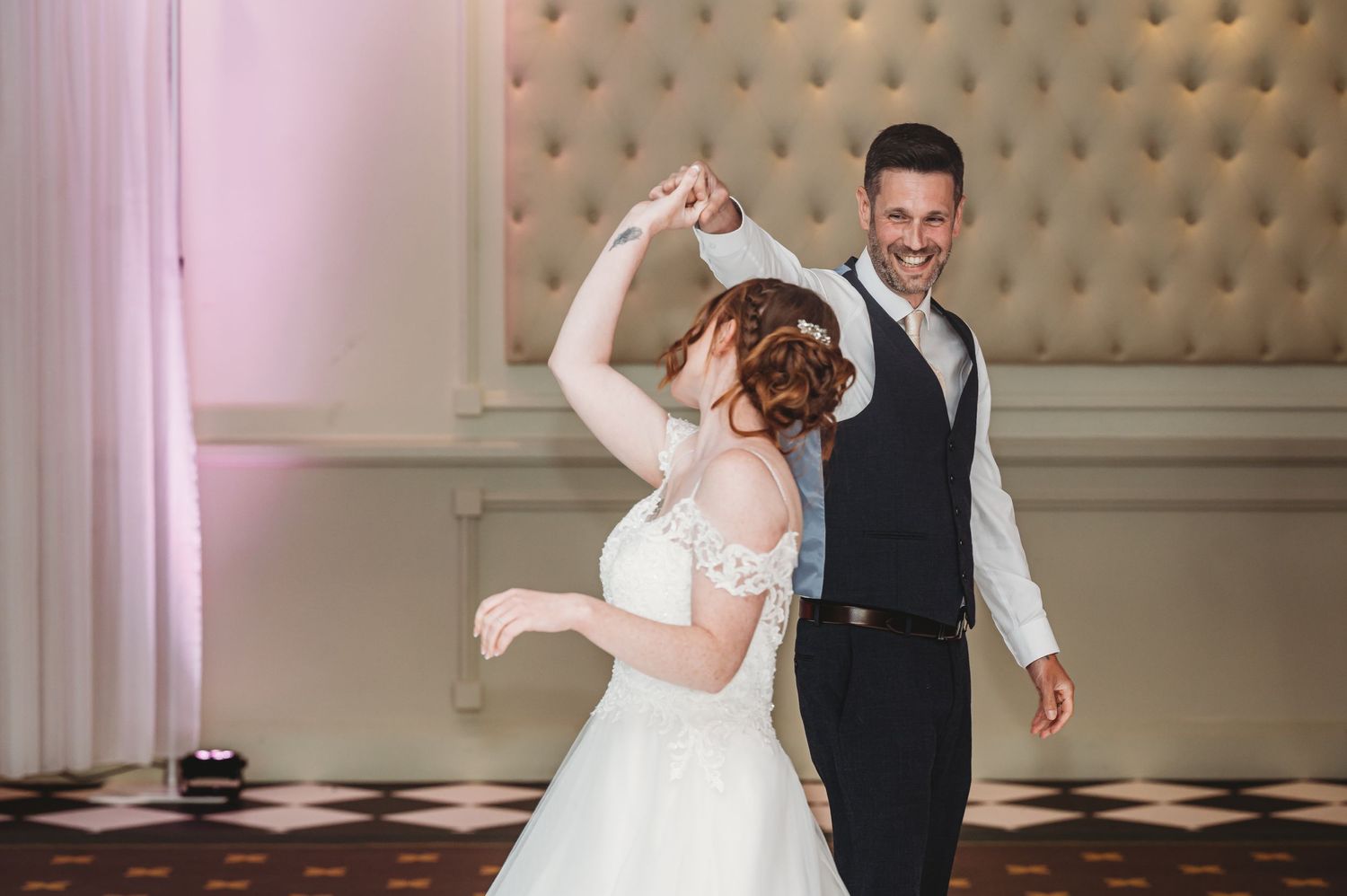 Two newlyweds sharing a joyful dance moment together in an elegant ballroom with tufted wall panels and checkered floor.