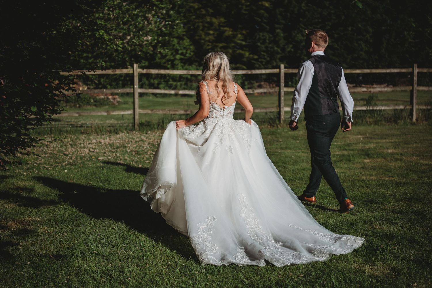 A couple walks across a sunlit lawn with a wooden fence, the bride's white gown trailing behind them.