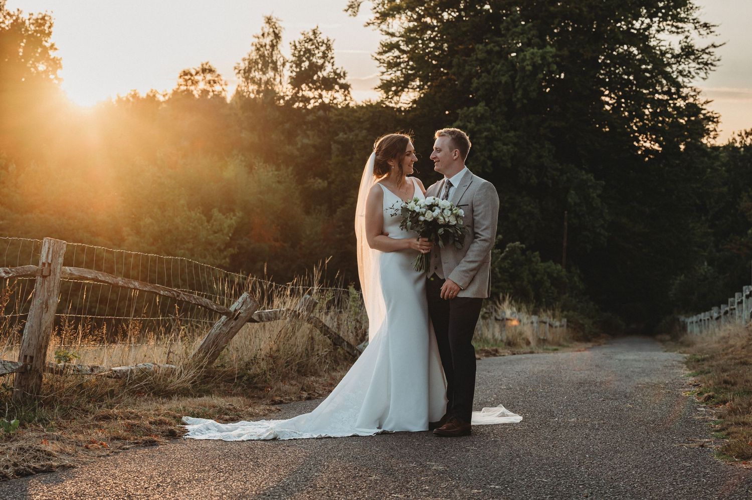A wedding couple embraces on a path during golden hour sunset with trees and natural scenery in the background.