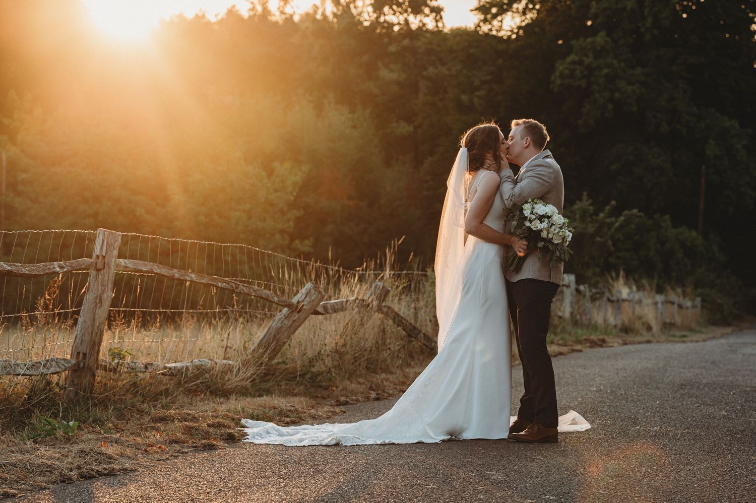 A romantic sunset wedding photo showing a couple embracing on a country road with a long white veil and bouquet.