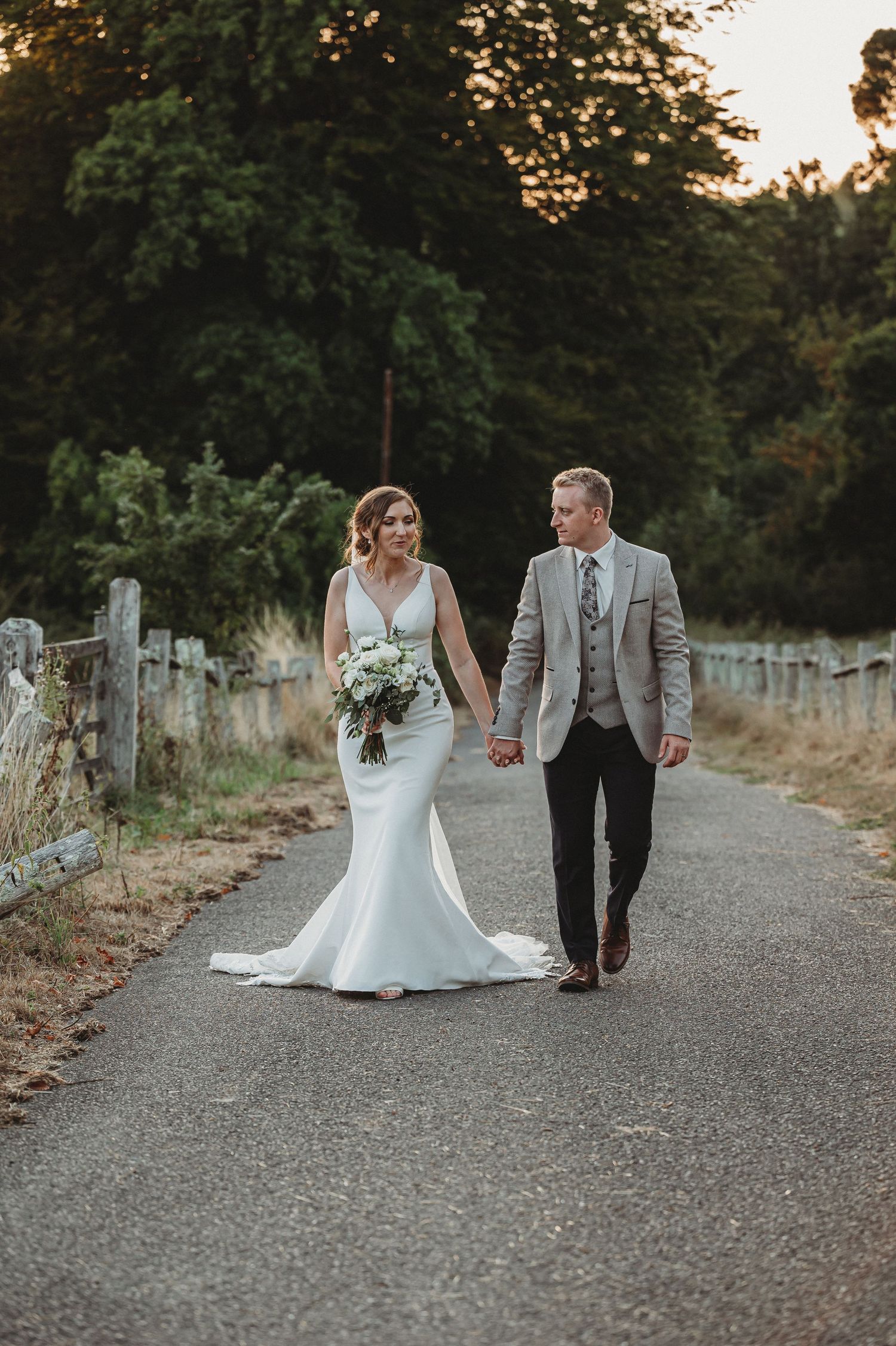 A couple walks hand in hand on a country path at sunset, dressed in elegant wedding attire with rustic fencing in background.