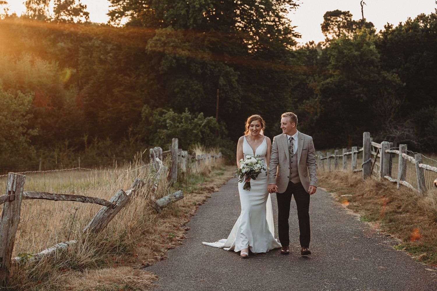 A couple walks hand in hand on a rural pathway during golden hour sunset, with rustic fence and trees in background.
