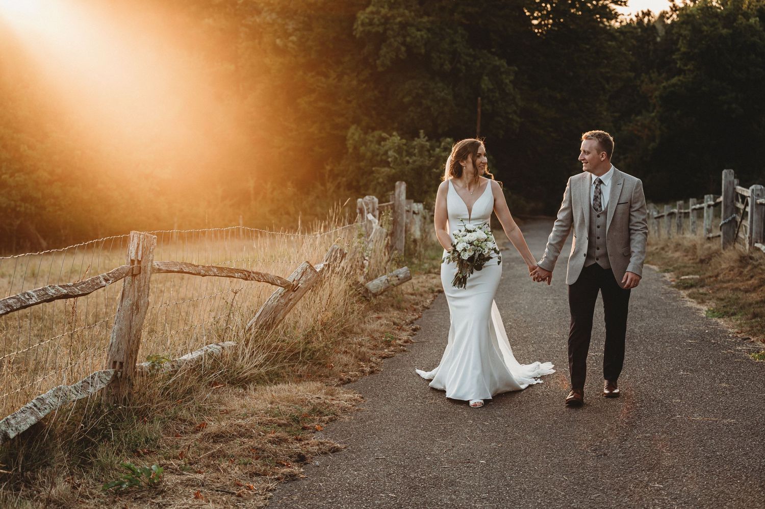 Newlyweds walk hand in hand on a country path at sunset with golden light streaming through trees.