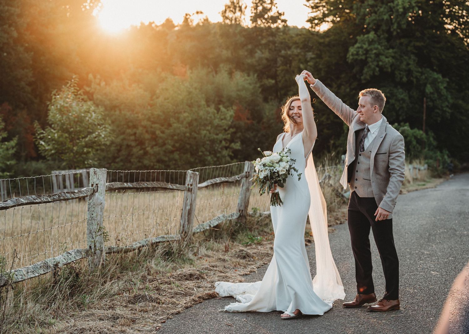 Newlywed couple dances together on a country road at golden hour with a rustic wooden fence in the background.