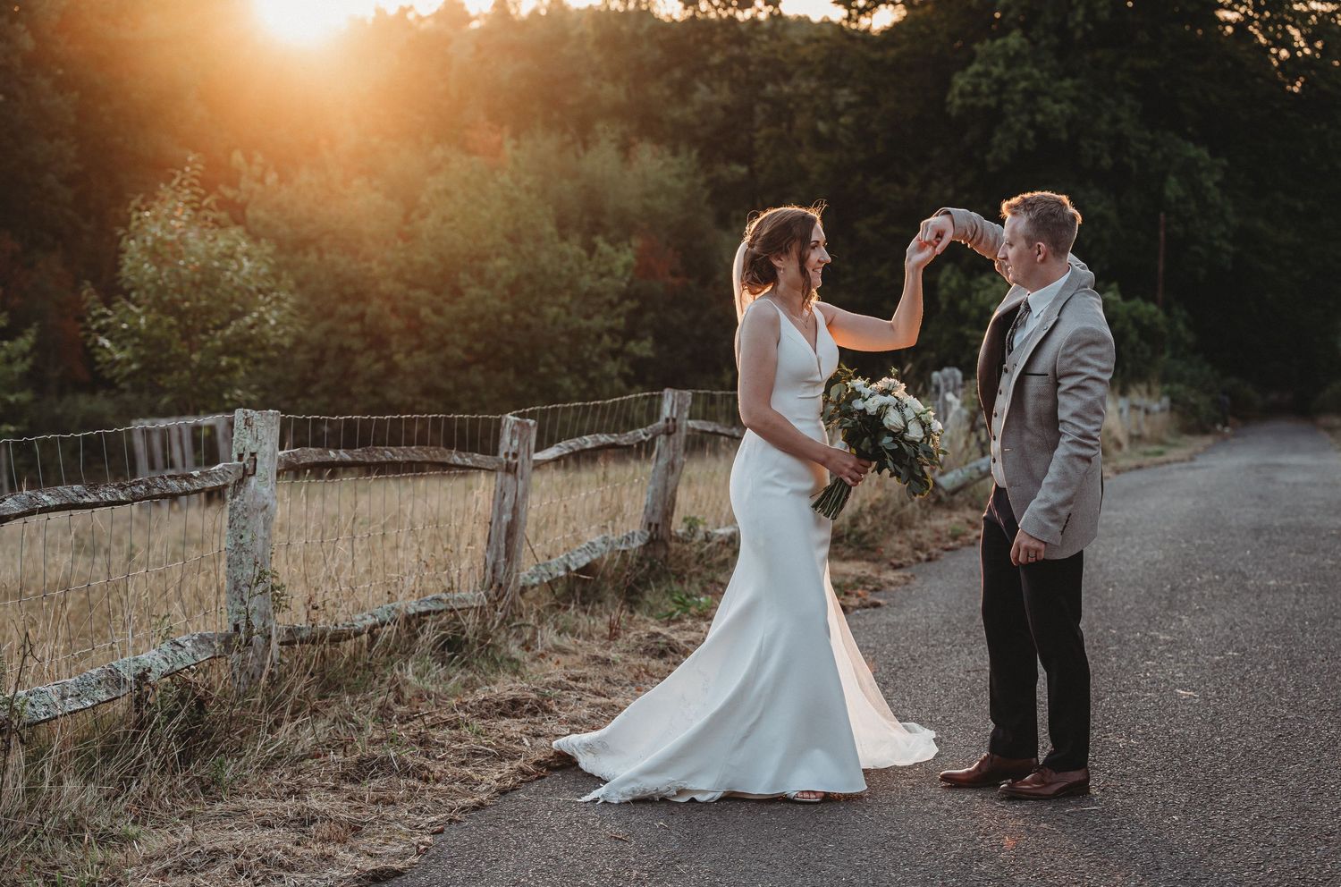 A couple shares a romantic sunset moment on a dirt path beside a rustic wooden fence during their wedding celebration.