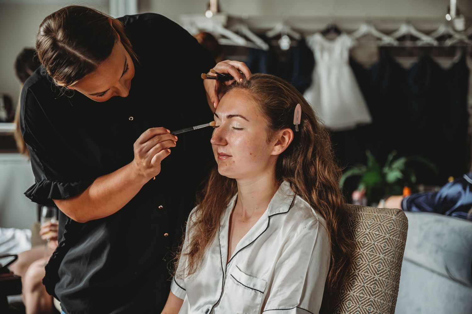 Makeup artist applies cosmetics to client wearing white silk robe during professional beauty session.