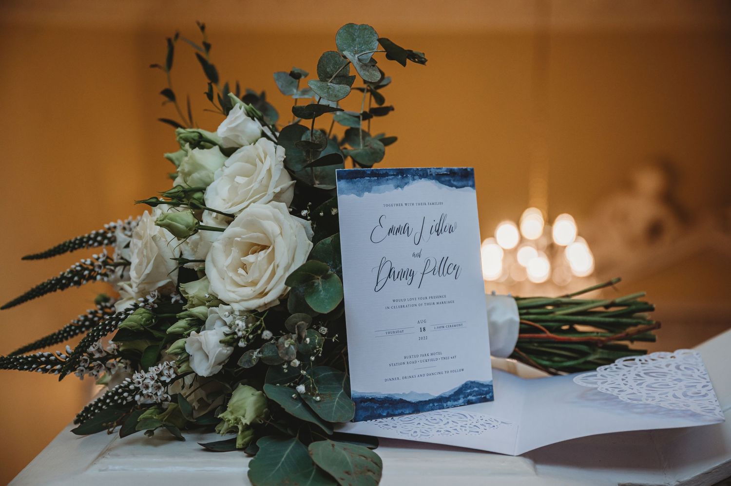 Wedding invitation displayed beside an elegant bouquet of white roses and eucalyptus with glowing candlelight in background.