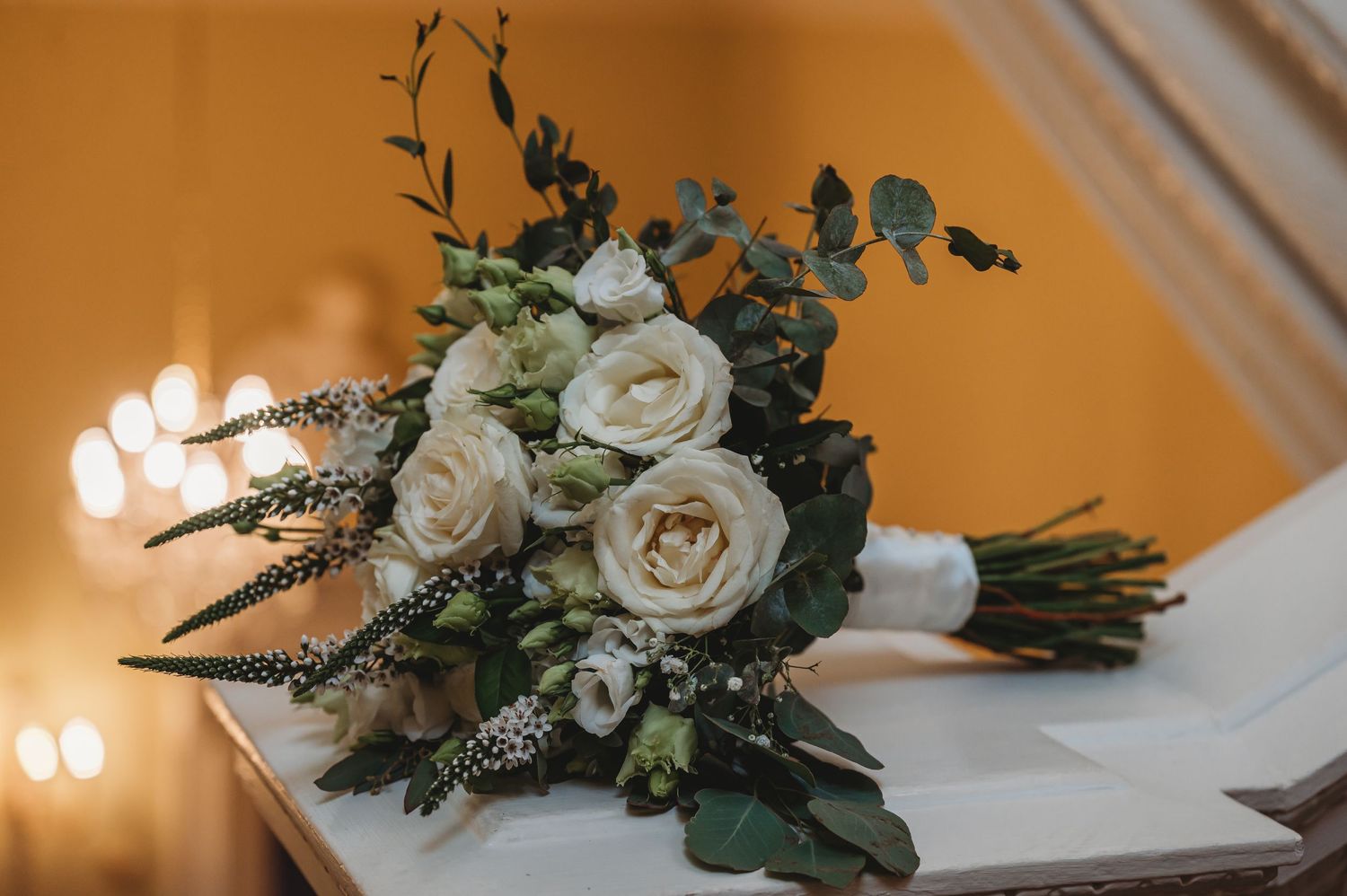 White rose bridal bouquet with eucalyptus greenery and delicate floral accents against an amber background.