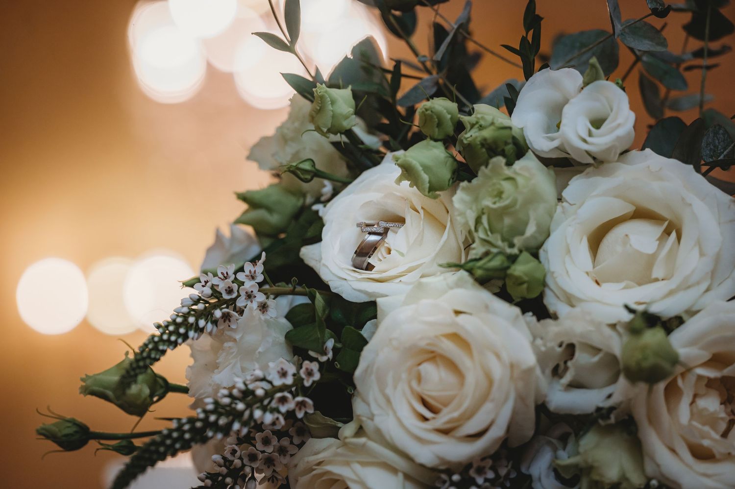 White roses in a soft-focus wedding bouquet with greenery and bokeh lights in the warm background.