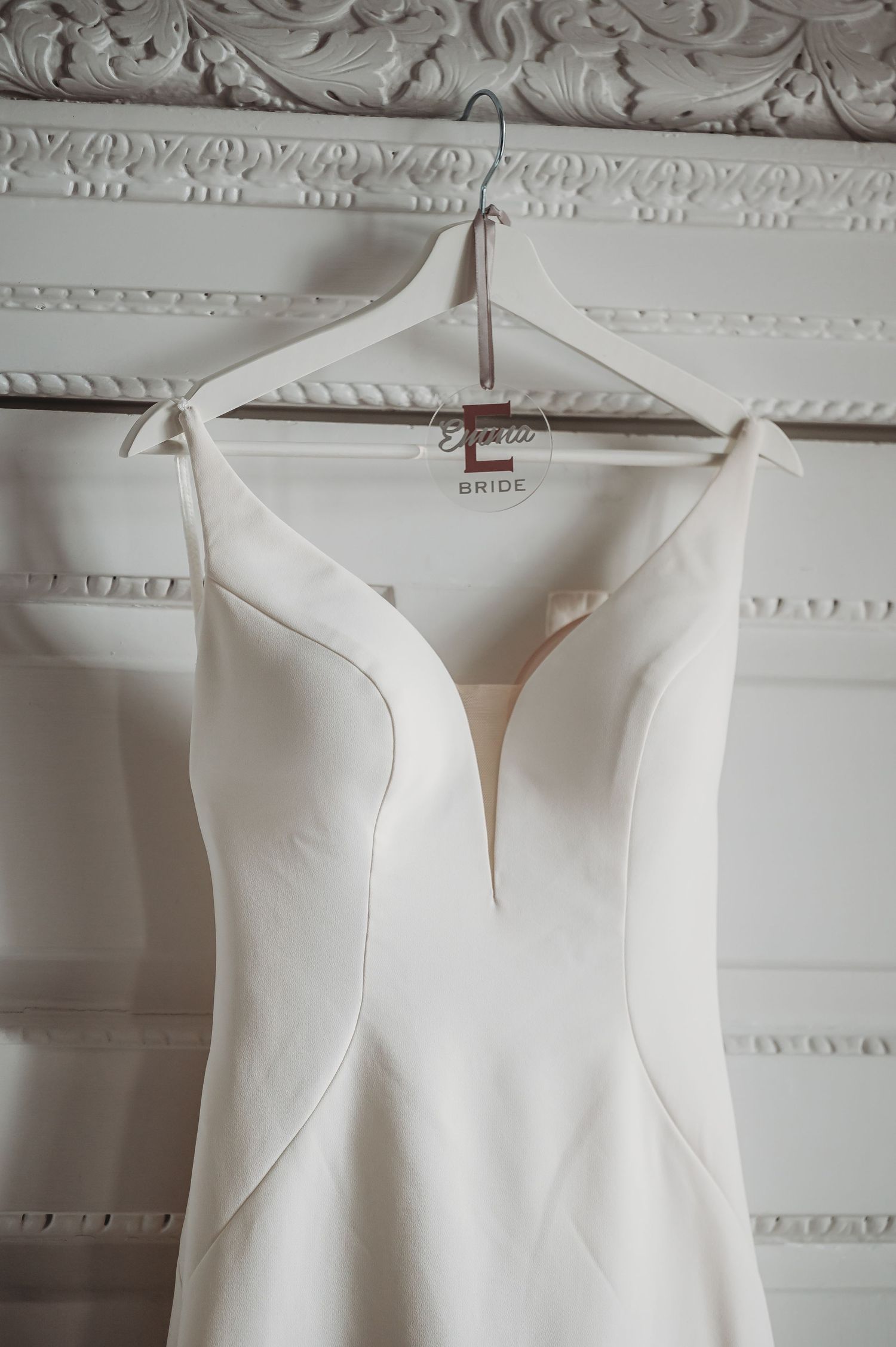 Elegant white wedding dress with plunging neckline and clean lines hangs on display against ornate molded wall.
