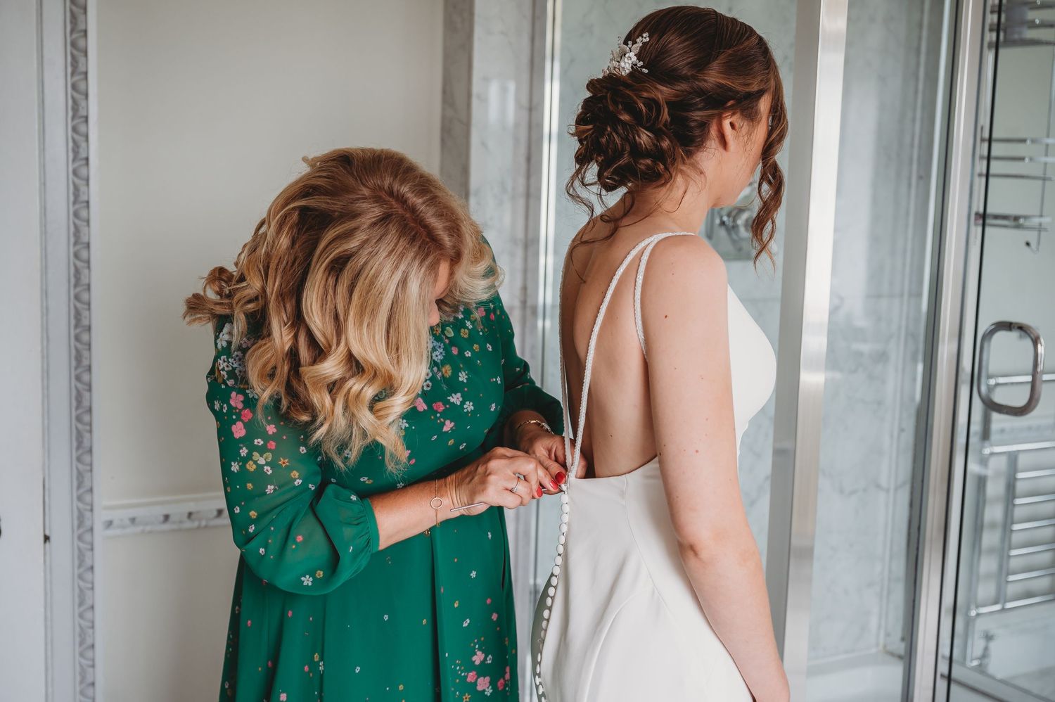 Two people stand together before a wedding, one wearing an elegant white dress while another assists in emerald green attire.