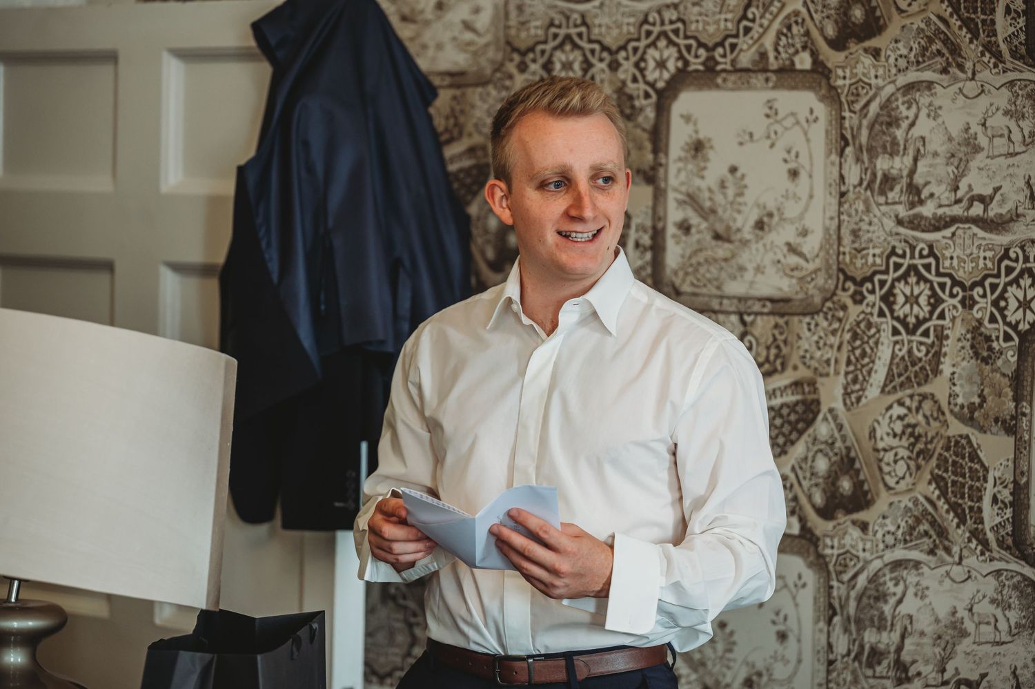 Someone preparing for a formal event smiles while holding papers in a room with decorative wallpaper.