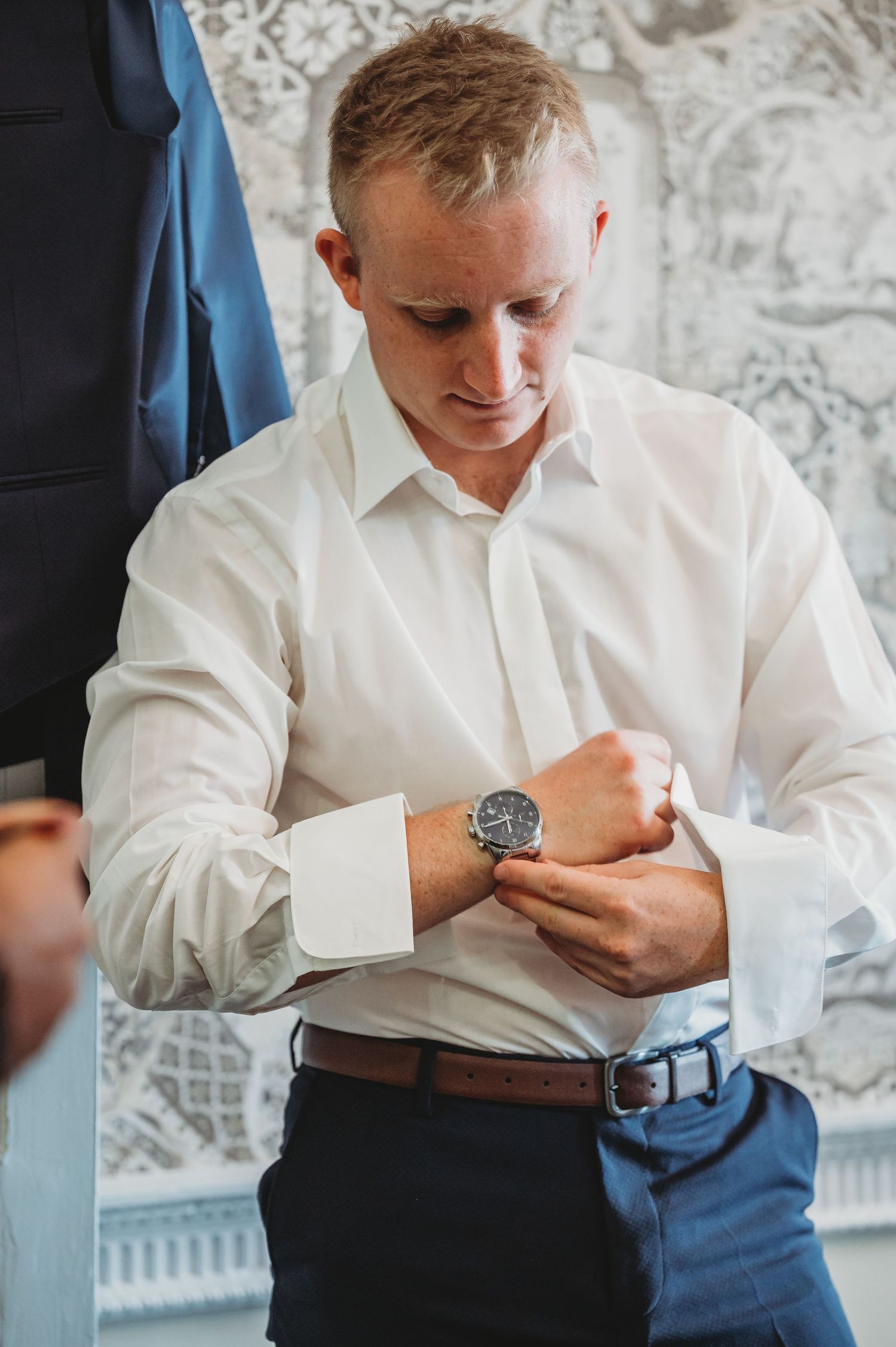 A person in a crisp white dress shirt and navy slacks adjusts a wristwatch while getting dressed.