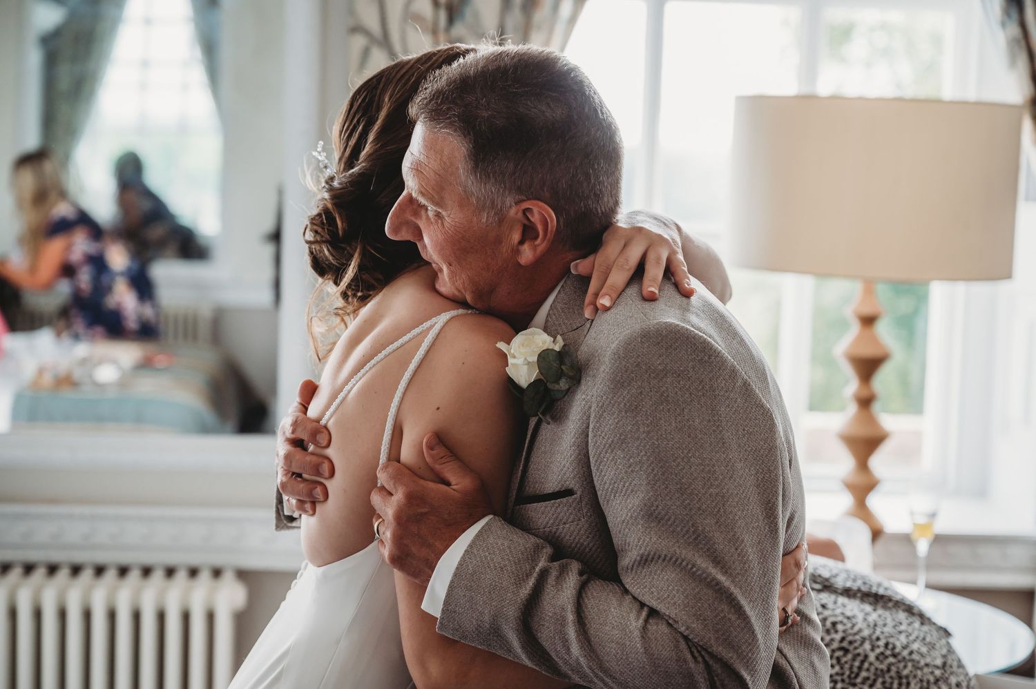 Two people share an emotional embrace during a wedding celebration, with soft lighting and a table lamp in the background.