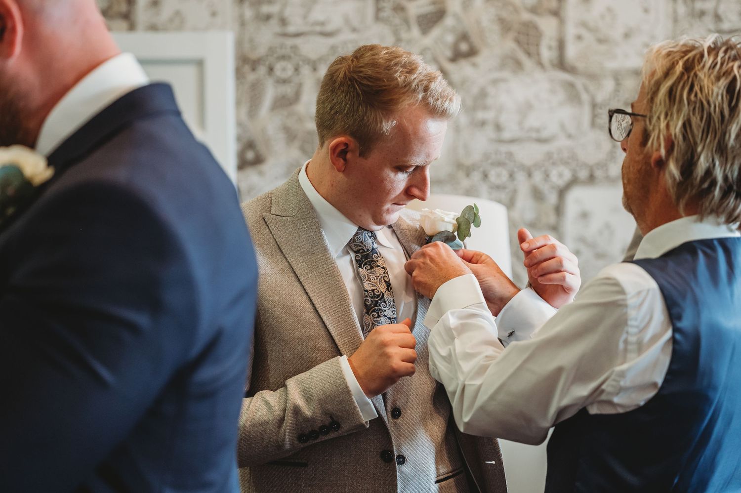 Wedding guests in formal attire inspect boutonnieres and accessories before a ceremony against a patterned wallpaper.