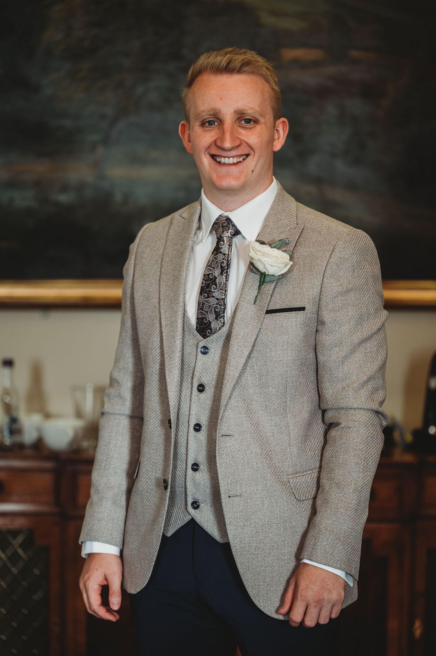 A person in a light gray three-piece suit with a patterned tie and white boutonniere smiles in an elegant indoor setting.