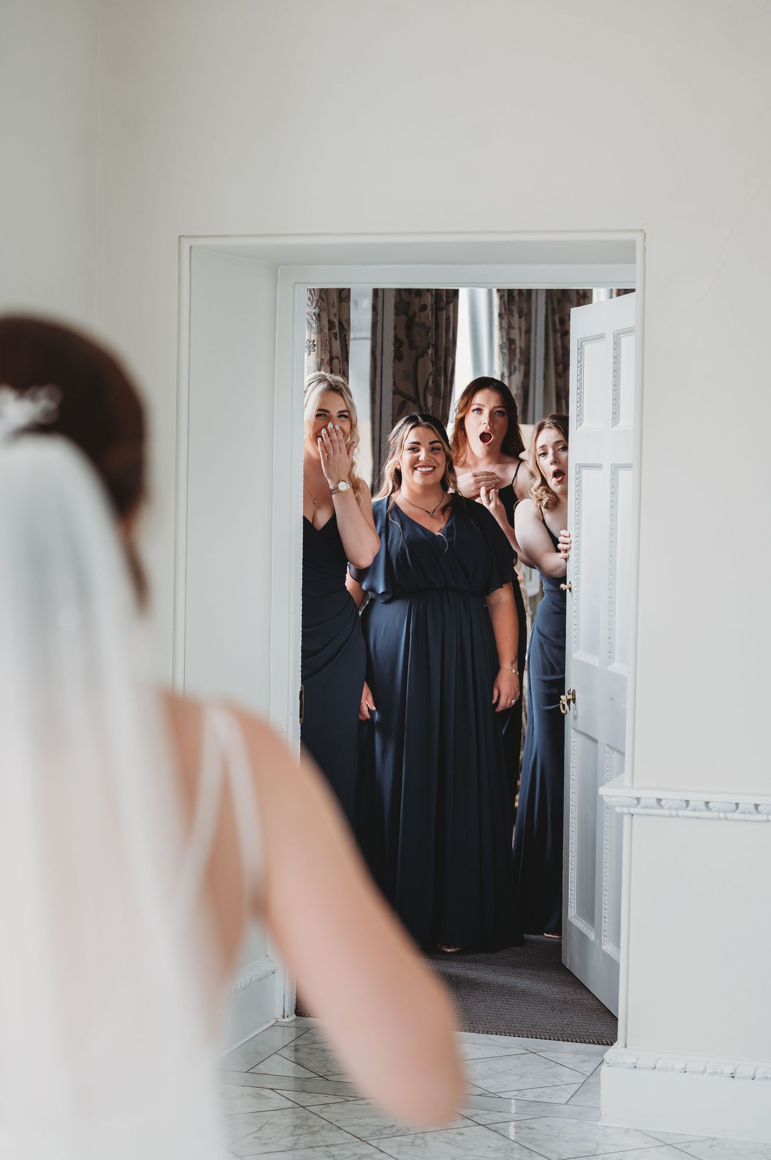 Bridesmaids in navy blue dresses react with joy and emotion while standing in a doorway during a wedding preparation.