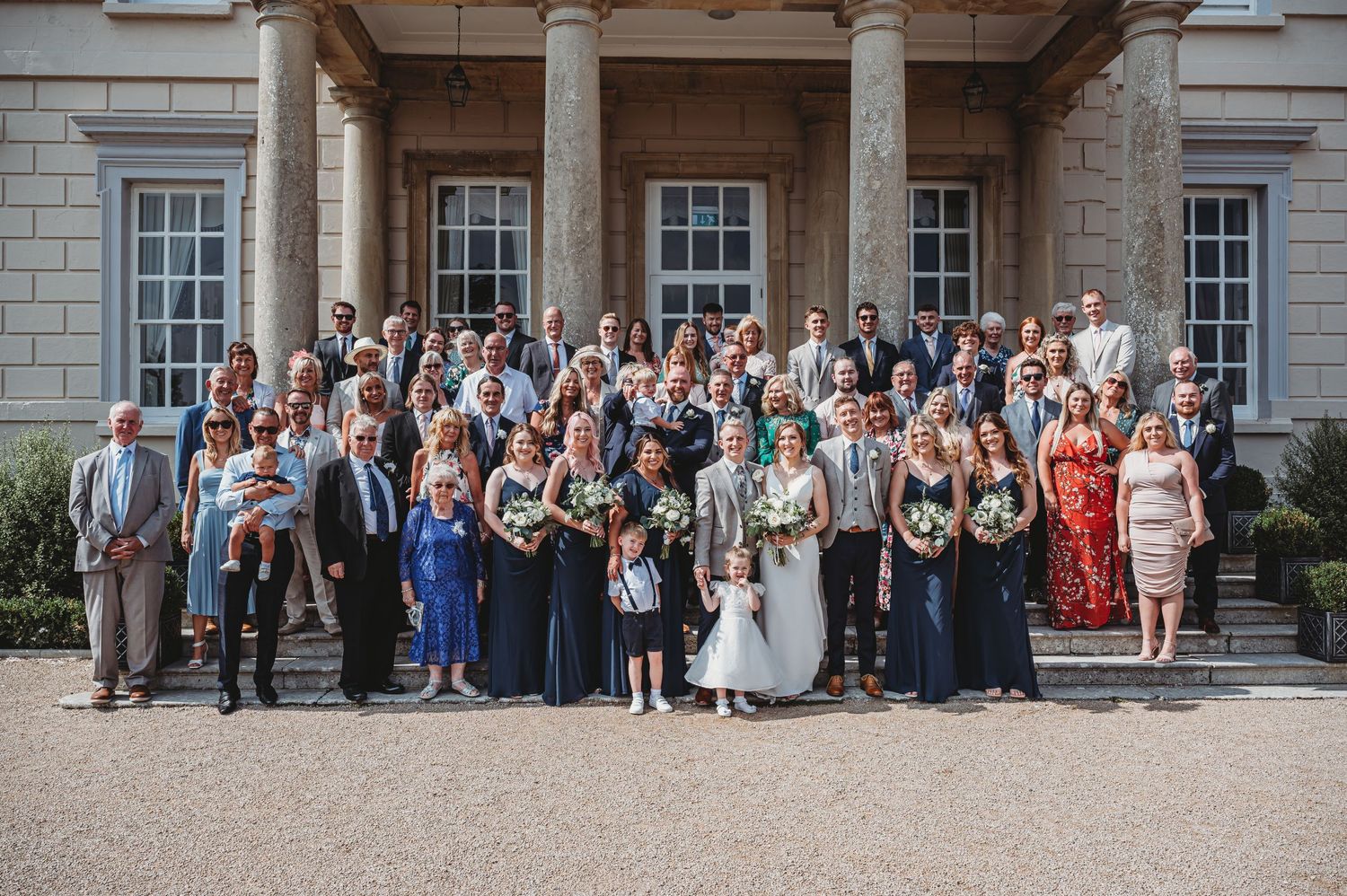 Large wedding party group photo gathered on the steps of an elegant stone building with columns.
