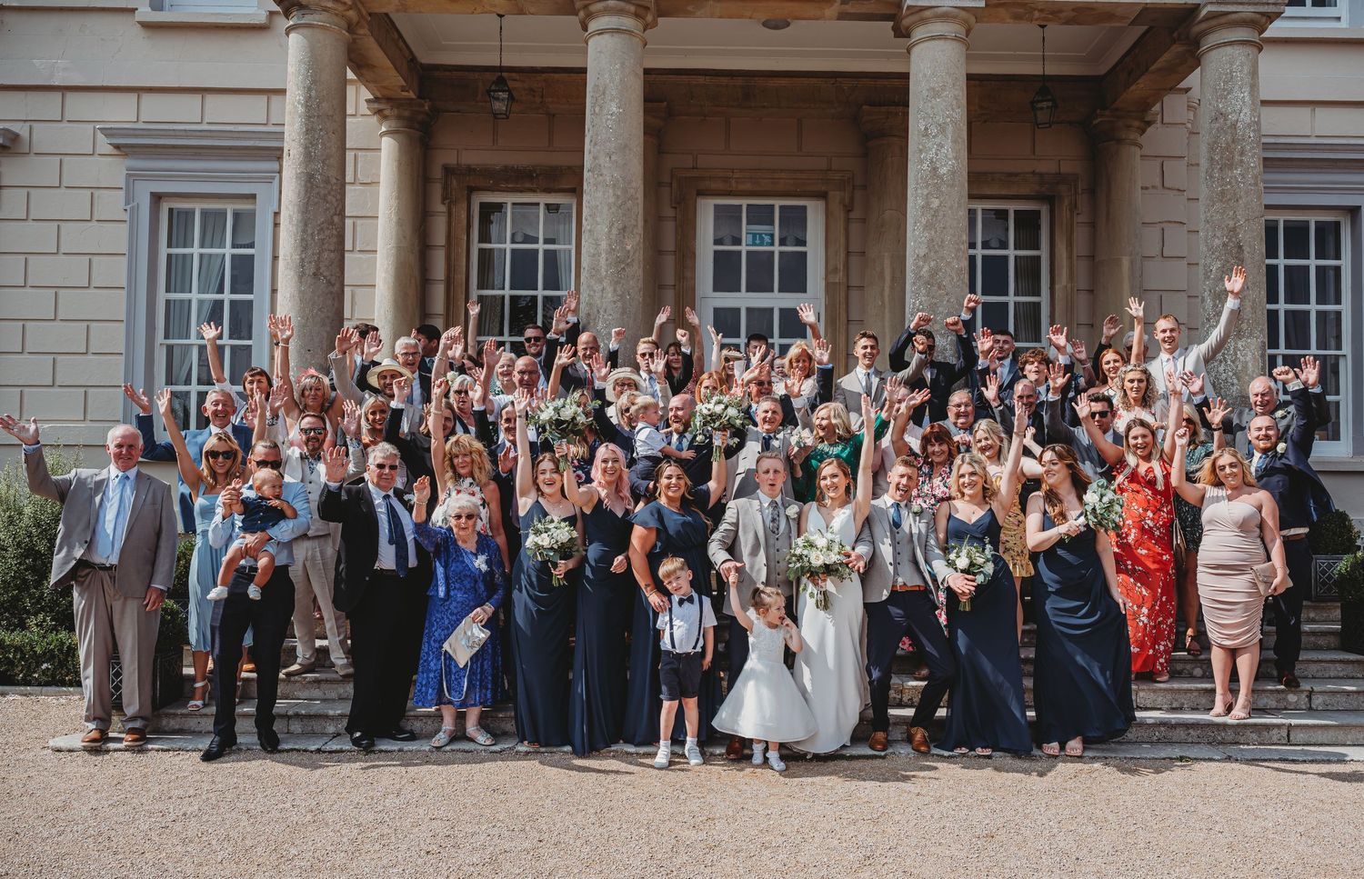 Large group of wedding guests gather on stairs outside grand historic building for celebratory photo.
