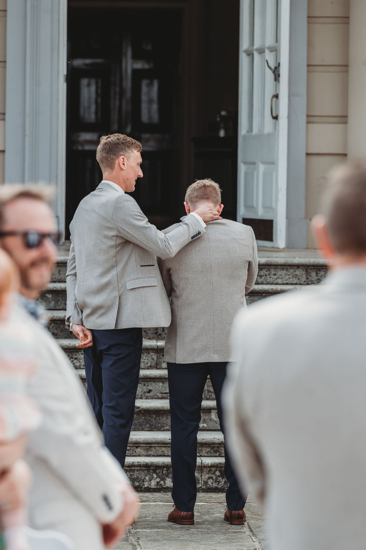 Two people in light gray suits stand on stone steps outside a building entrance during what appears to be a formal event.