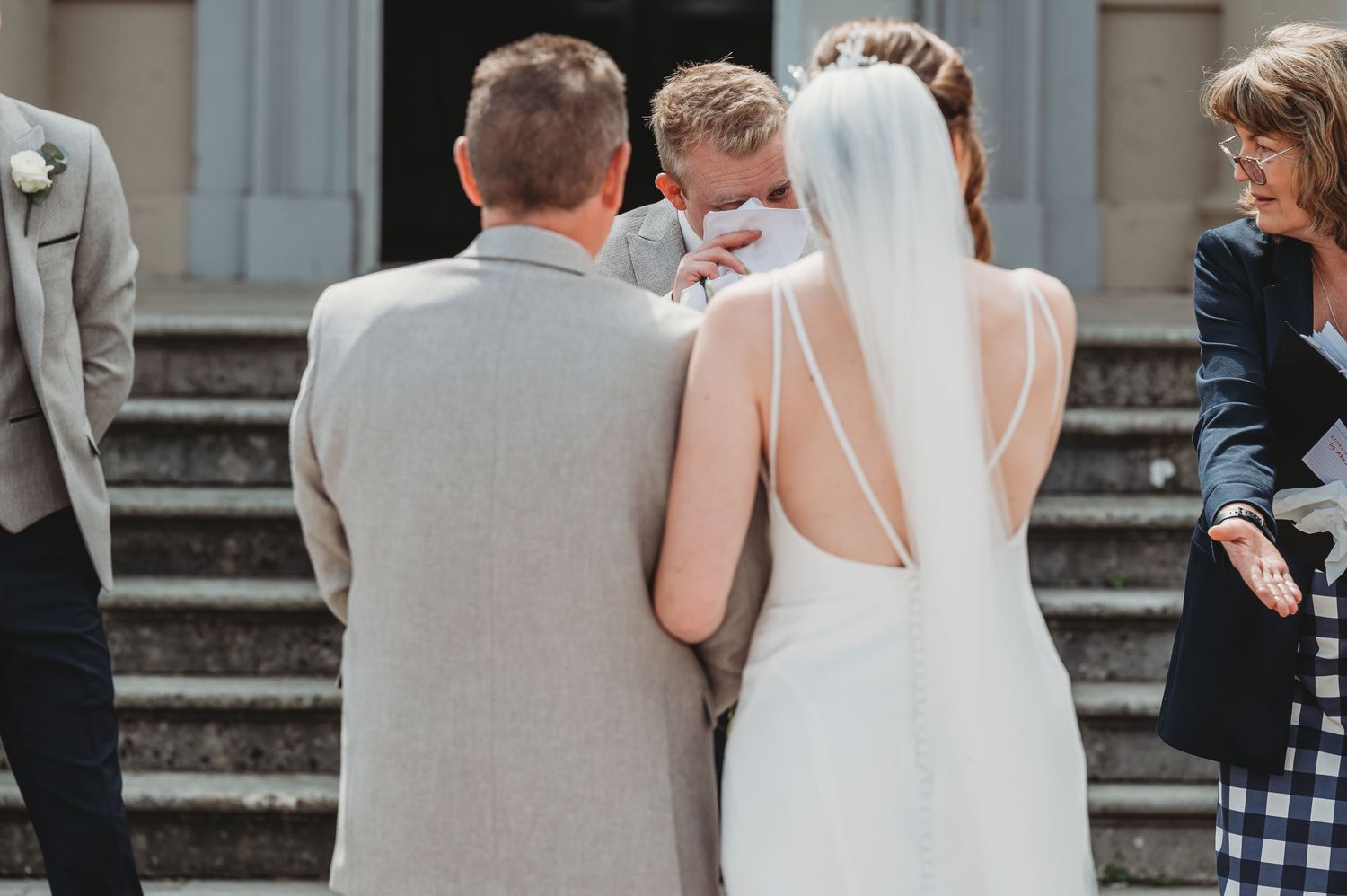 A bride in a white dress stands with guests on stone steps at an outdoor wedding venue.