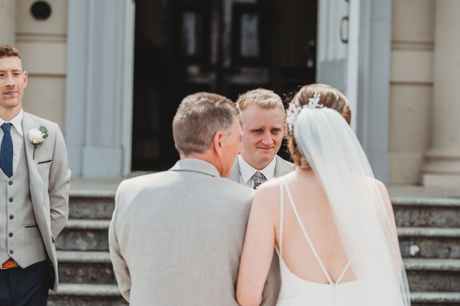 Wedding ceremony on building steps with bride in backless dress and guests in formal attire gathered outdoors.