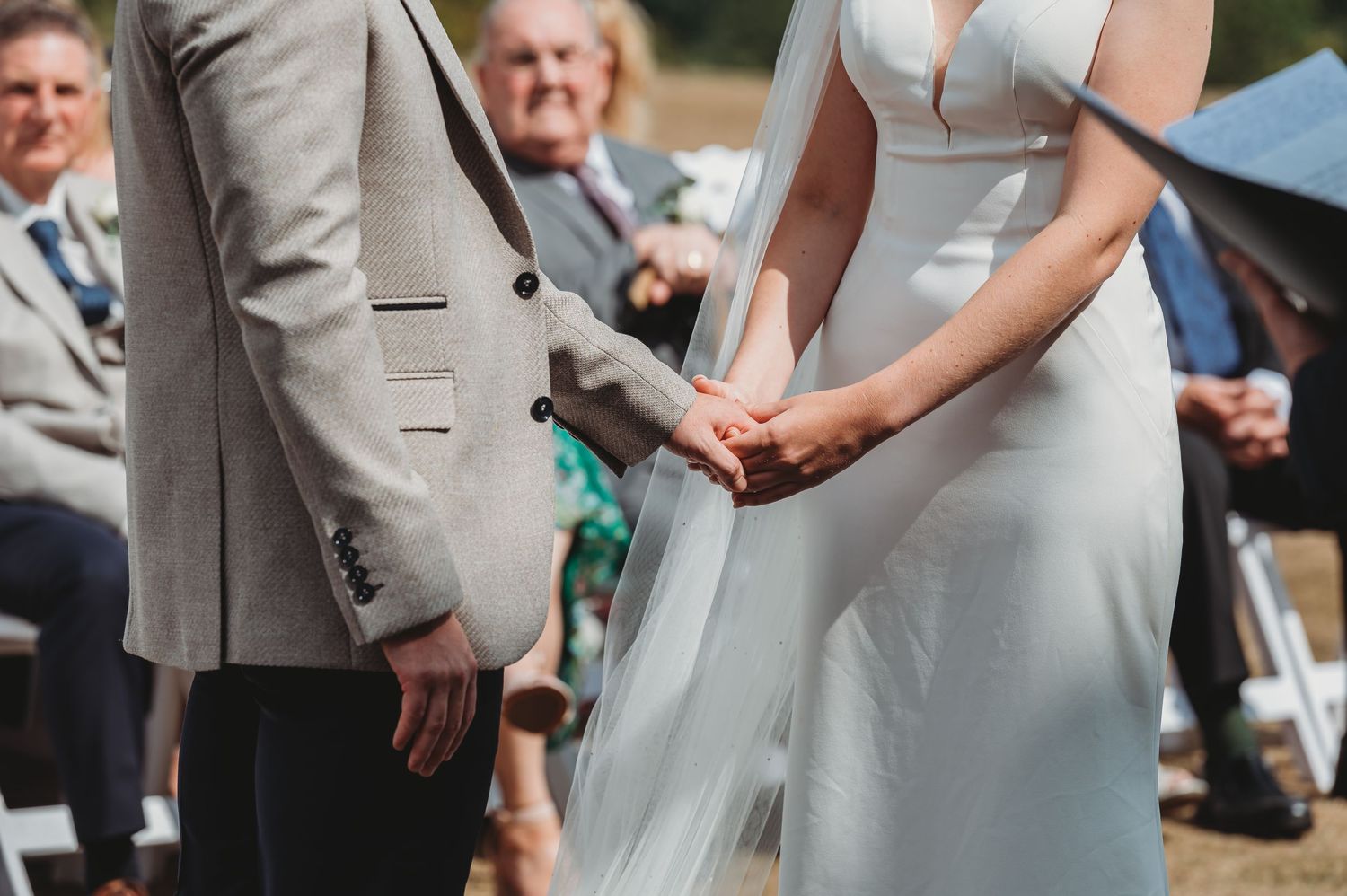 Close-up shot of a couple holding hands during an outdoor wedding ceremony in elegant gray and white attire.