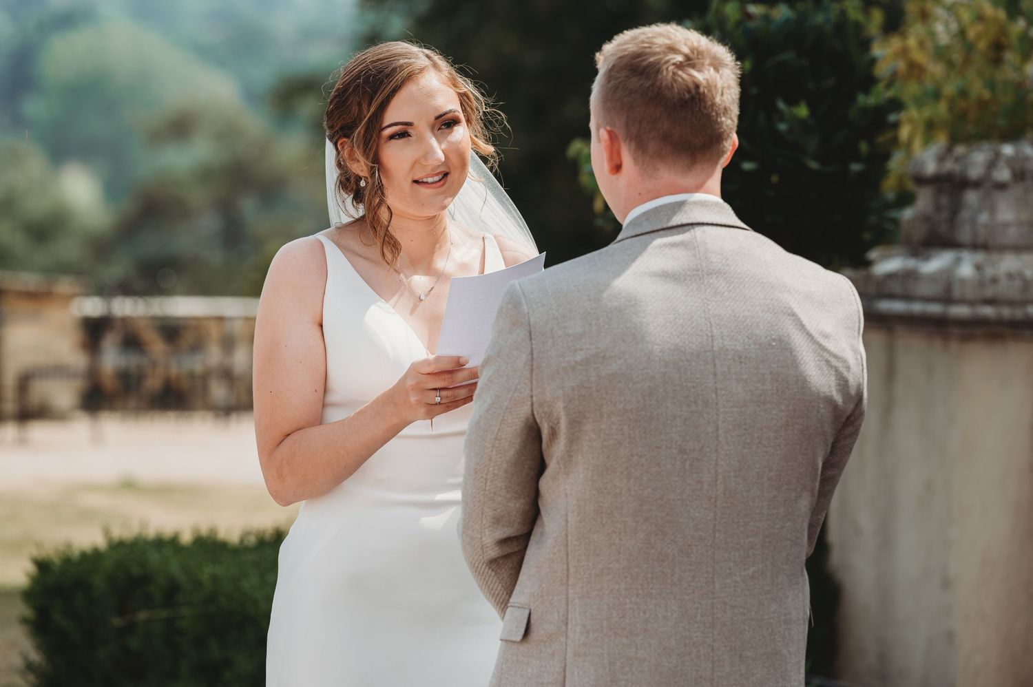 A bride in a white wedding dress reads vows during an outdoor ceremony while facing the groom in a grey suit.