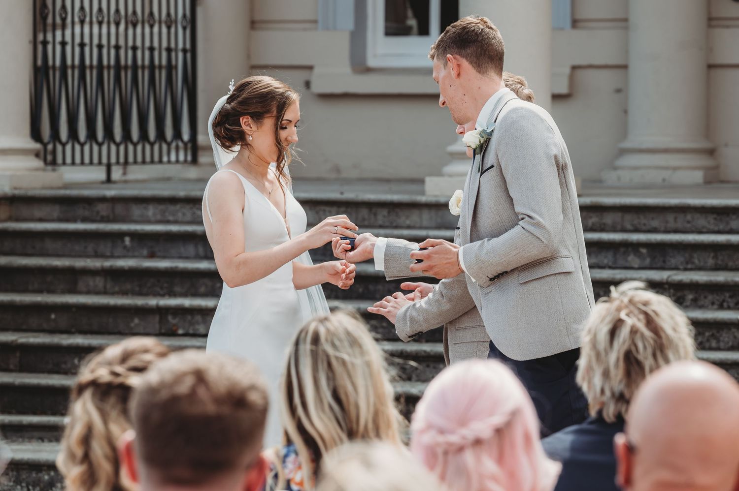 A joyful outdoor wedding ceremony exchange of rings on stone steps with seated guests watching below.