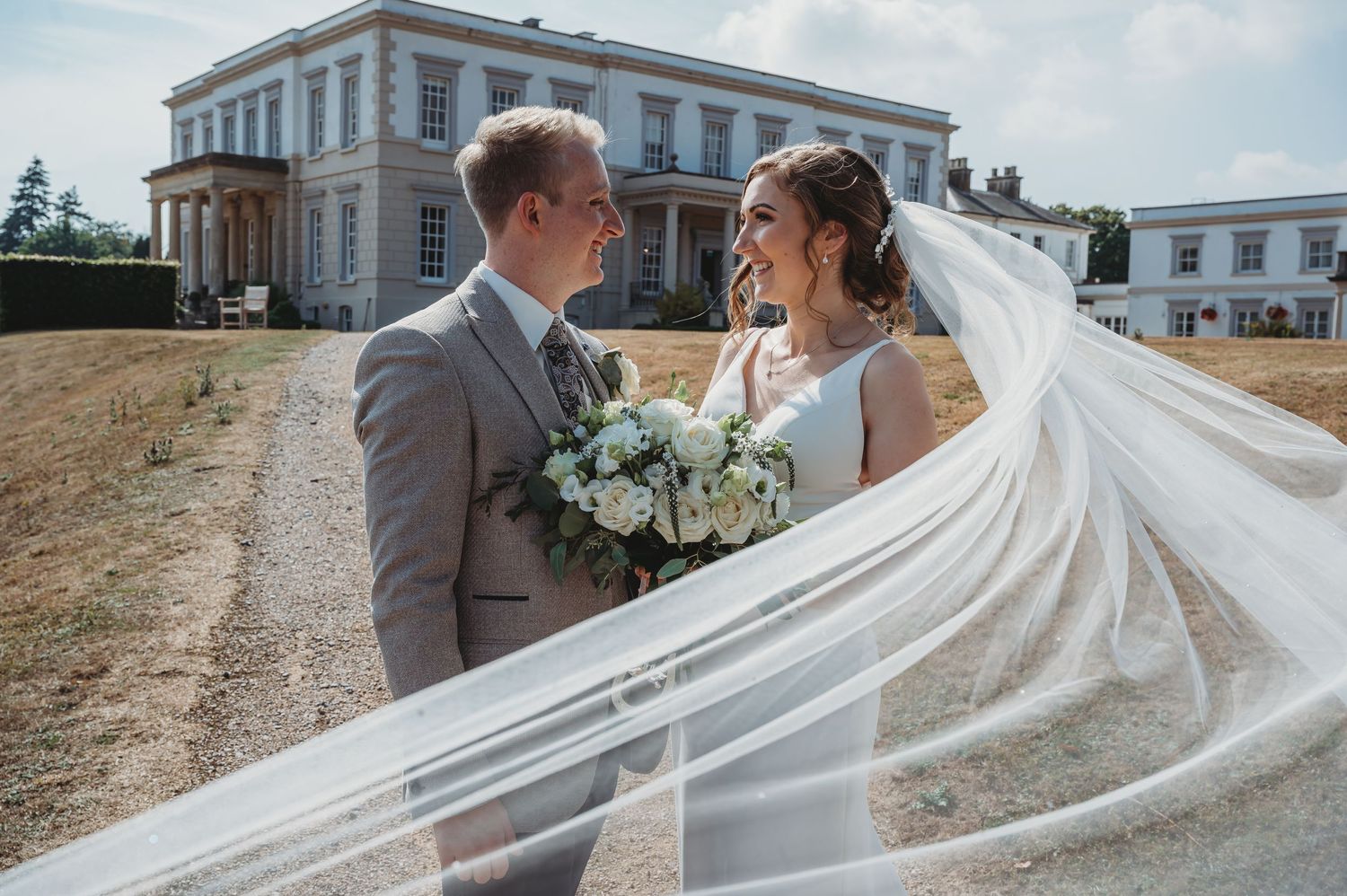 A wedding couple shares an intimate moment outside a stately mansion while a long flowing veil catches the light.