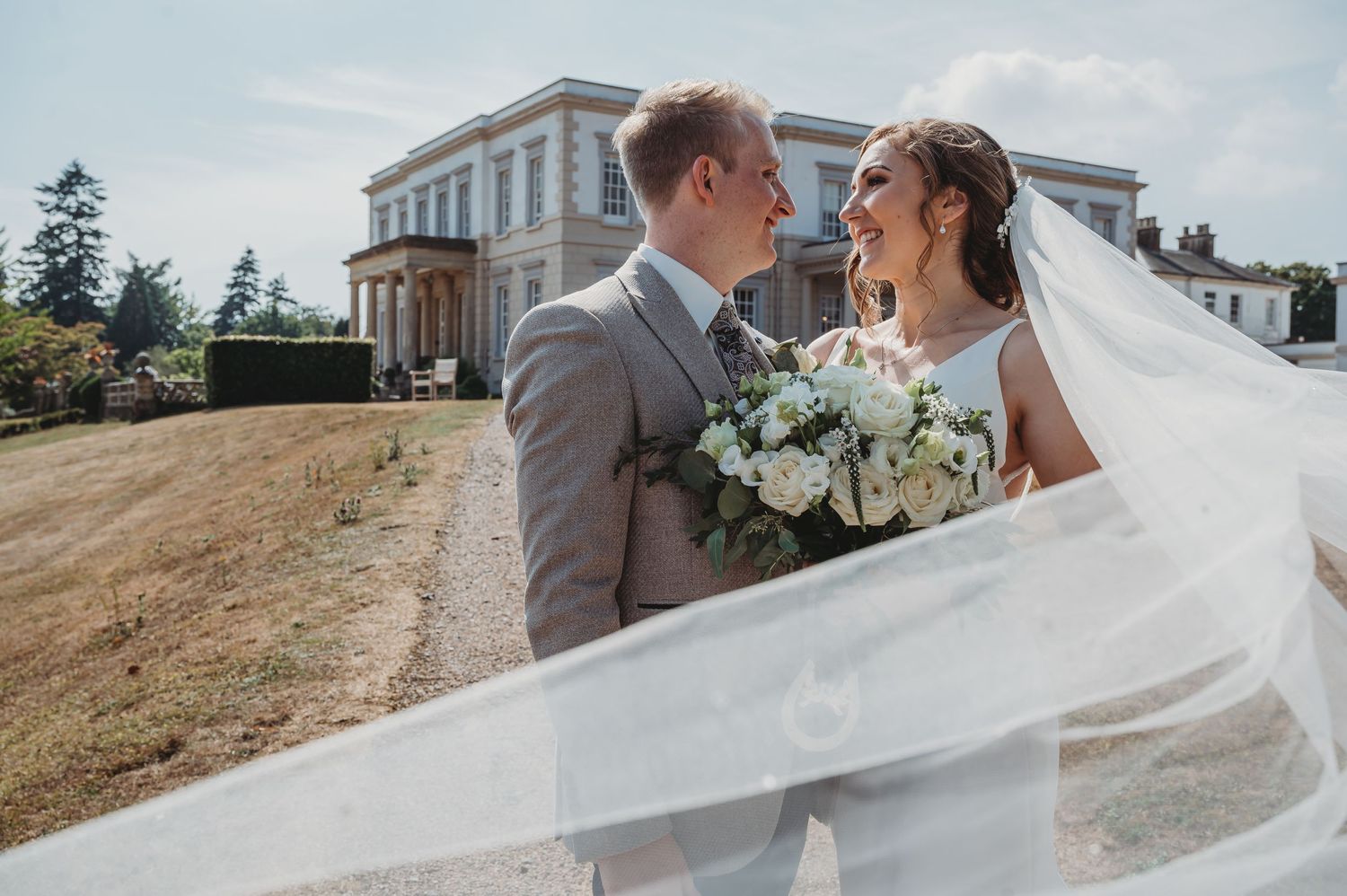 A wedding couple shares a tender moment in front of an elegant white mansion as the bride's veil flows in the breeze.
