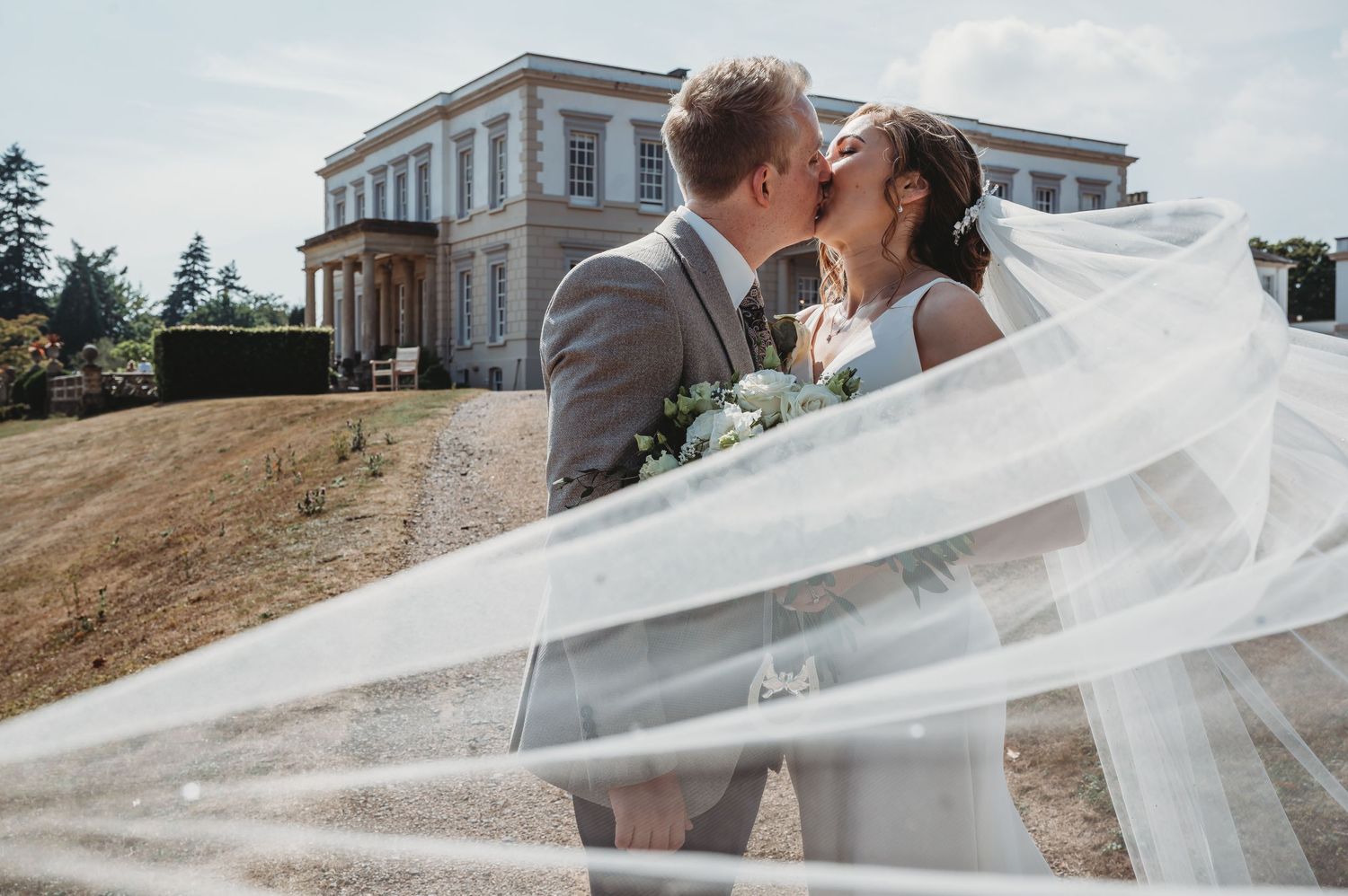 A couple shares a romantic kiss on their wedding day, with a flowing veil and elegant mansion in the background.