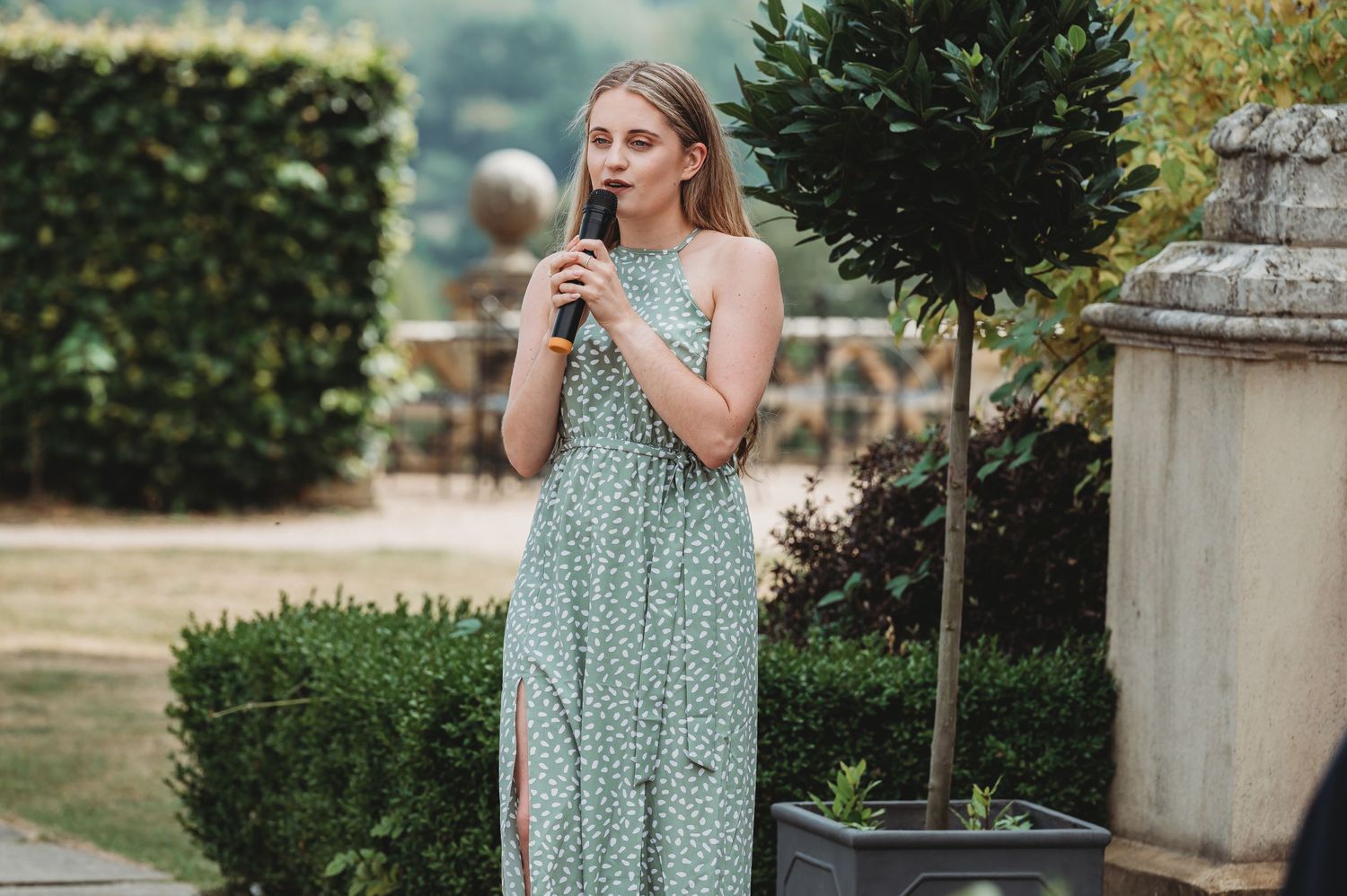 A person in a mint green floral dress stands outdoors near manicured topiary and stone planters on a sunny day.