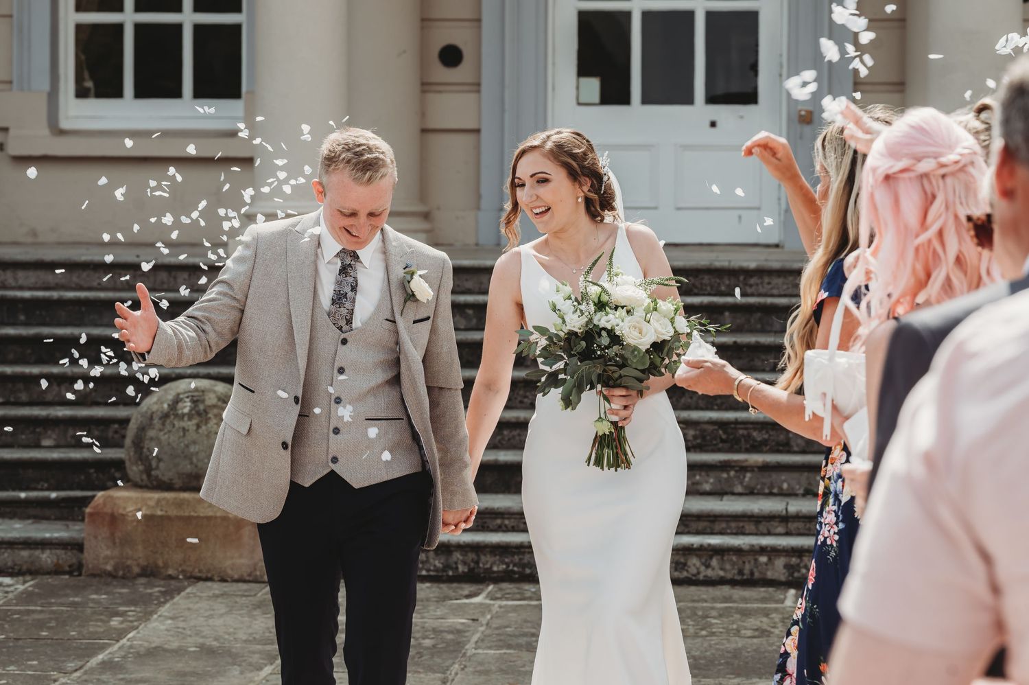A joyful wedding couple walks down stone steps while guests shower them with confetti in celebration.