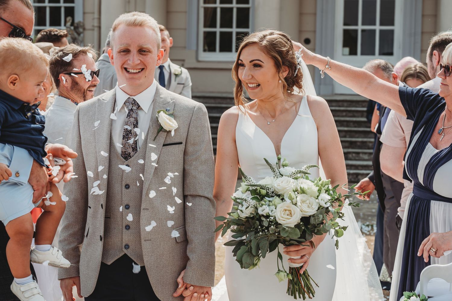 A wedding couple smiles as guests throw flower petals in front of a white building during their recessional.