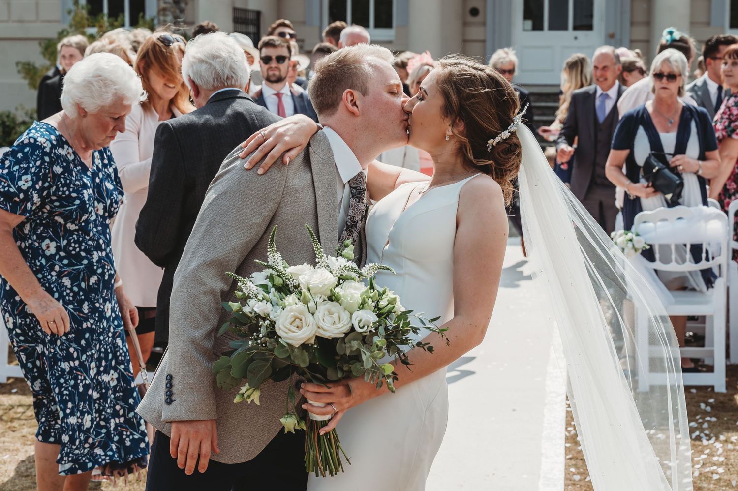 A romantic outdoor wedding ceremony with a newlywed couple embracing in a kiss while holding a white floral bouquet.