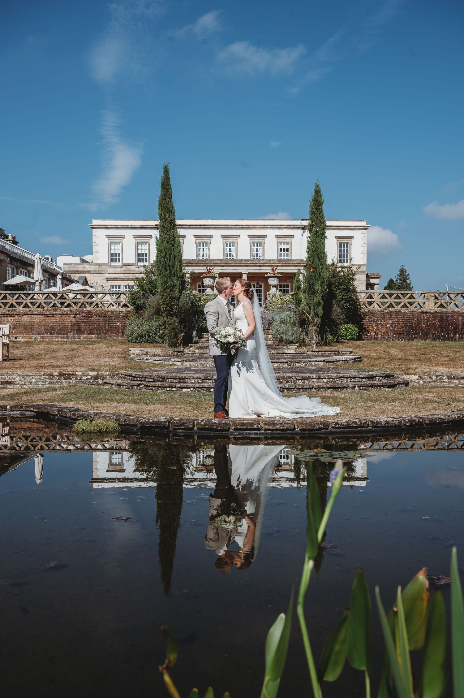 A romantic couple poses at a Mediterranean villa with tall cypress trees and a reflective pool creating a mirror image.