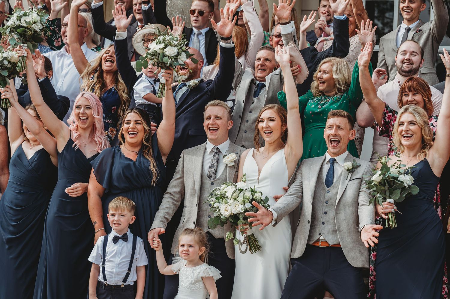 Wedding party in navy dresses and gray suits celebrating joyfully together with raised arms and bouquets.
